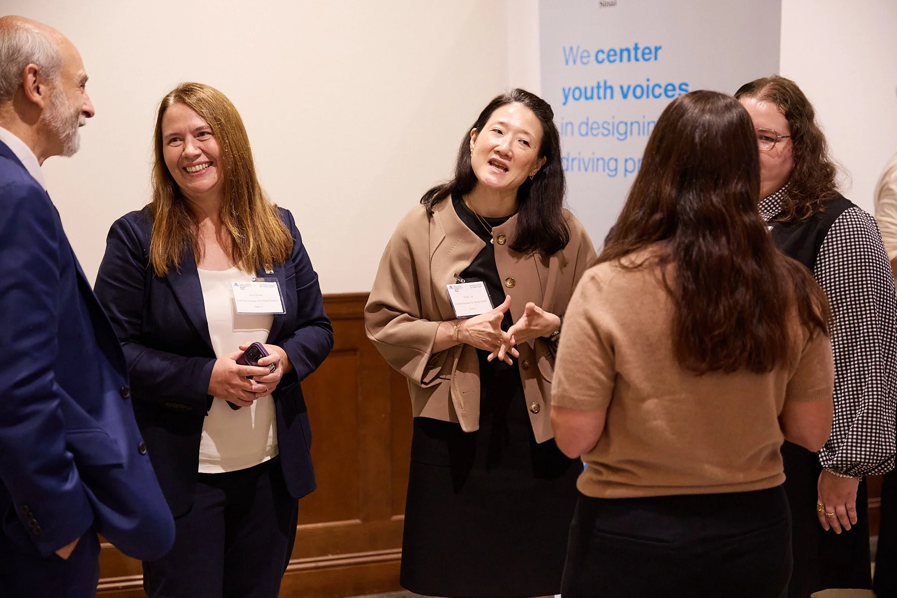 Group of five women and one man having a conversation at a professional event, with a presentation screen in the background that reads "We center youth voices in designing driving p".