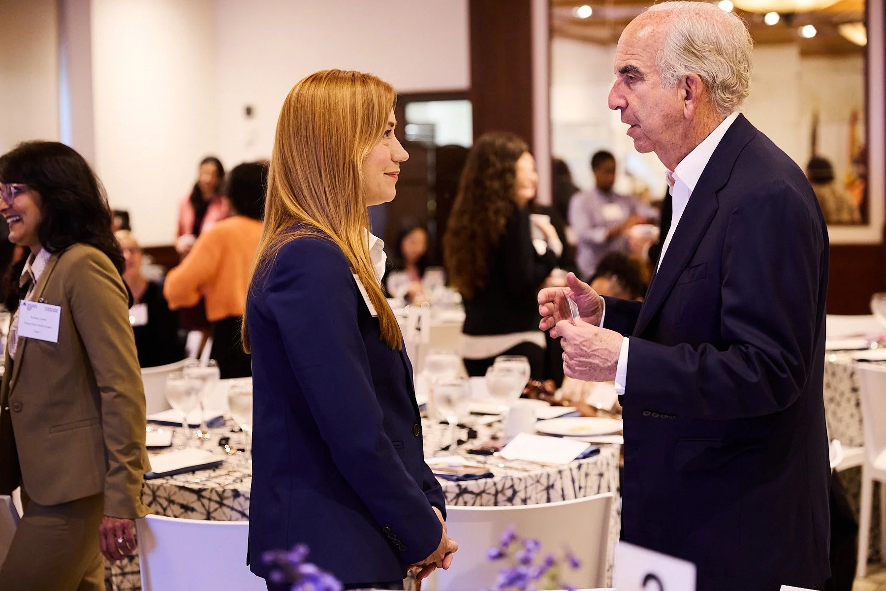 A young woman and an older man in conversation at a professional event, with other attendees and tables in the background.