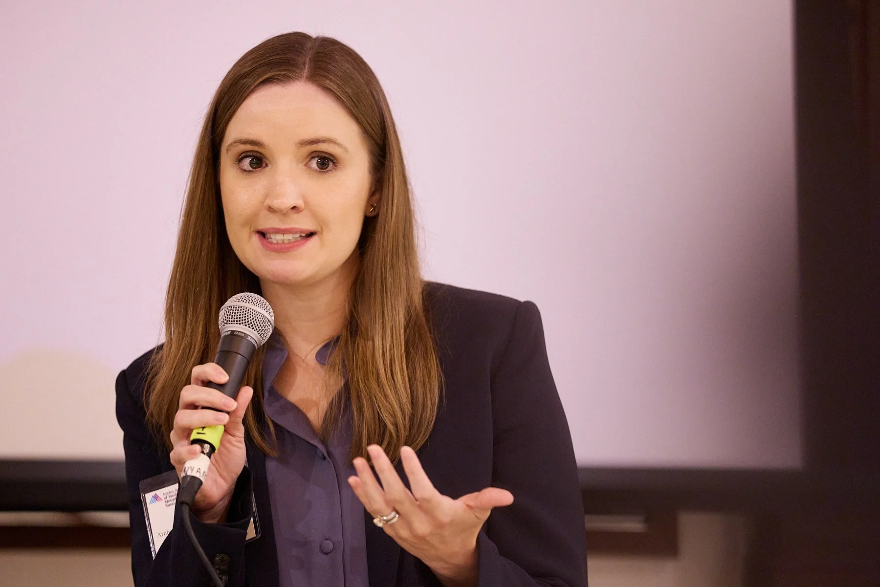 A woman with long brown hair wearing a dark blazer and purple shirt speaking into a microphone during a presentation or speech.
