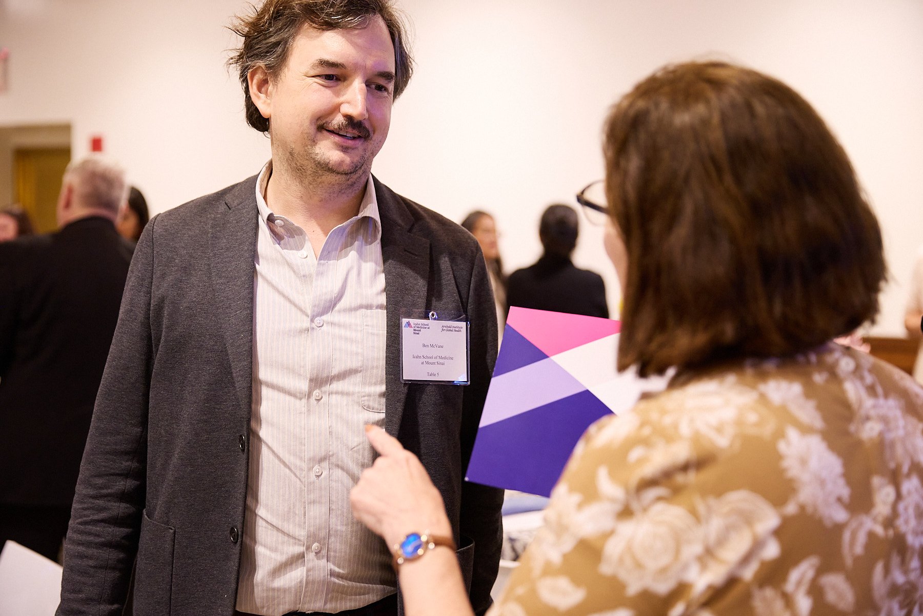 A man in a gray blazer talking to a woman holding a brochure at a professional event.