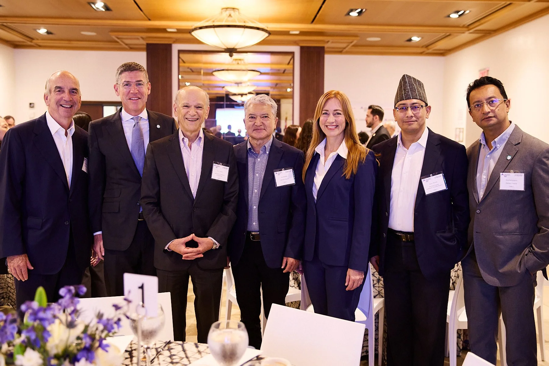 Group of seven professionally dressed people in suits standing together at a conference or formal event in a well-lit room with wood-paneled ceiling and reception tables.