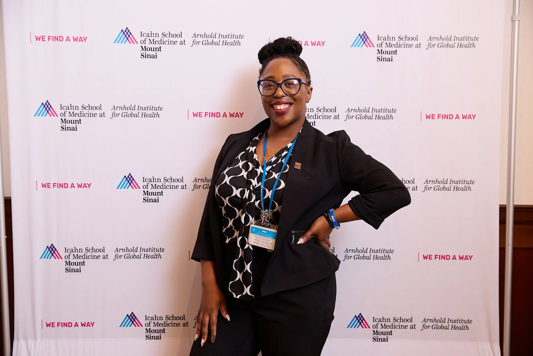 A smiling woman standing in front of a backdrop with logos and text related to Icahn School of Medicine at Mount Sinai and Arnold Institute for Global Health. She is wearing glasses, a black blazer, and a patterned black and white blouse, with confer