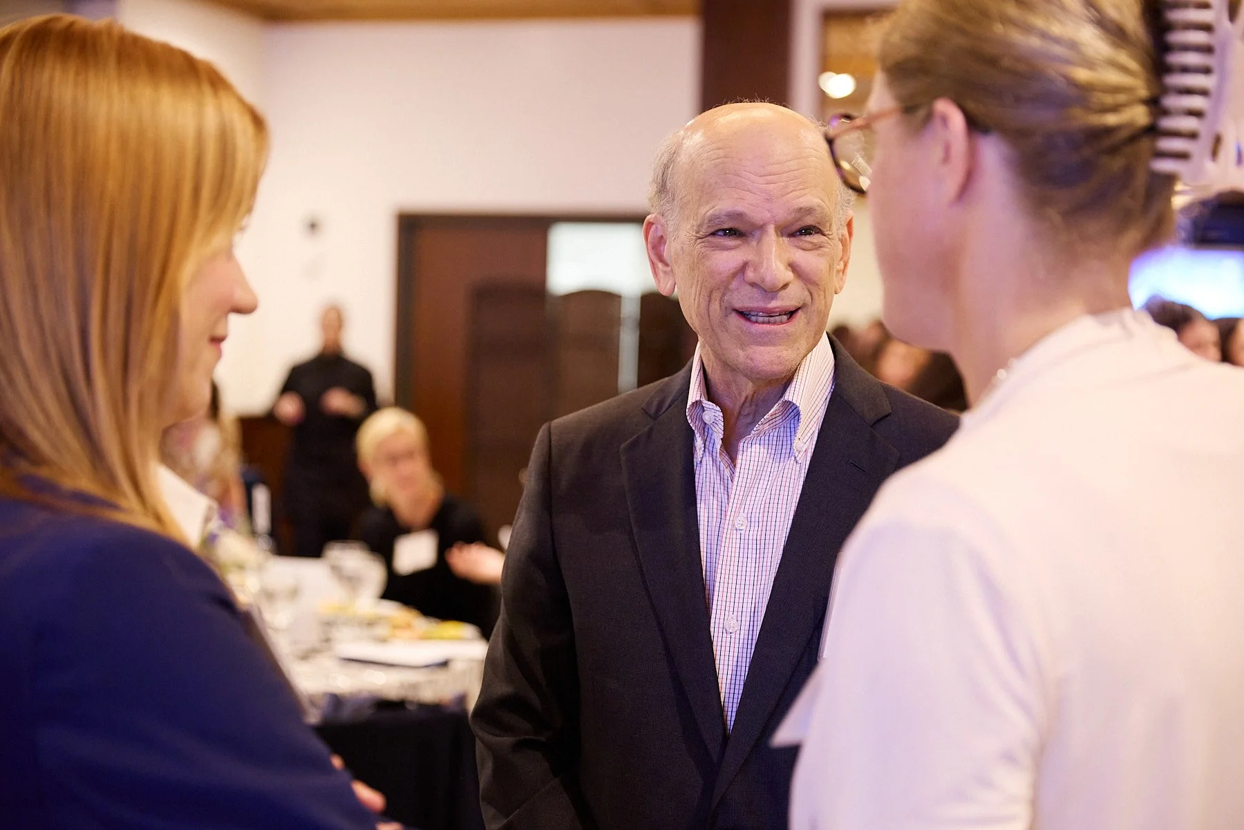 An older man in a suit smiling and talking to two women at an indoor event. One woman has red hair and the other has blonde hair tied back.