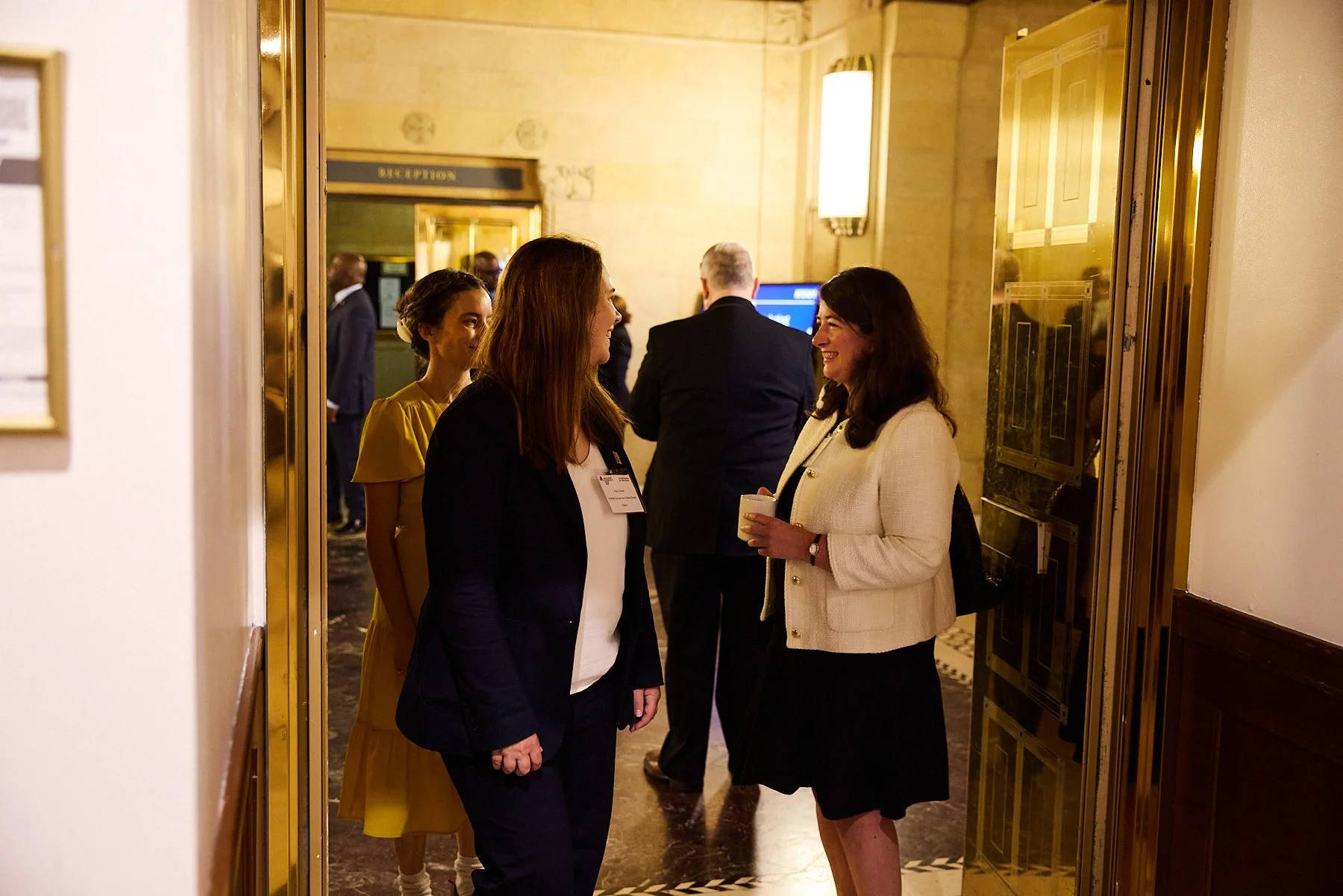 Two women engaged in conversation at a social event, with other people in the background, in a warmly lit, elegant venue.