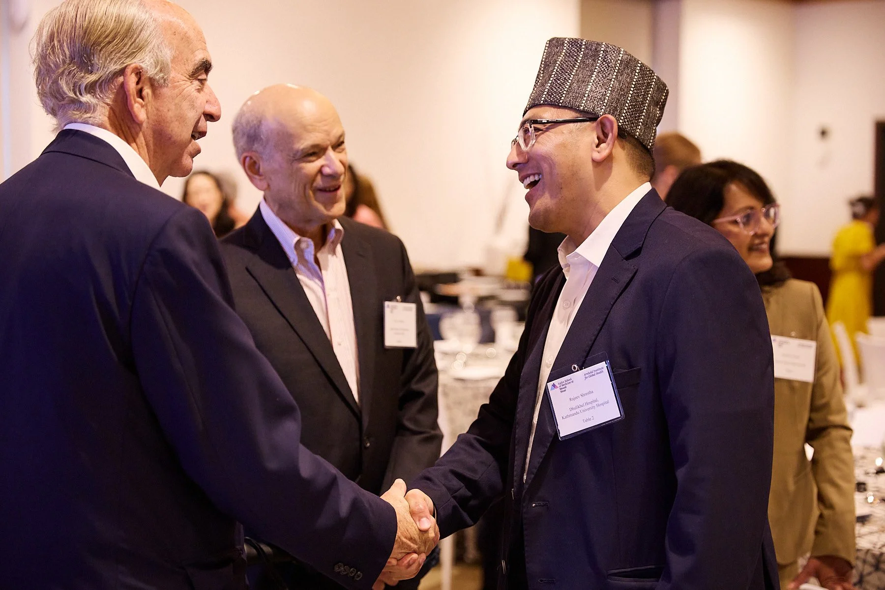 Three men in suits shaking hands at a professional event, with other attendees in the background.