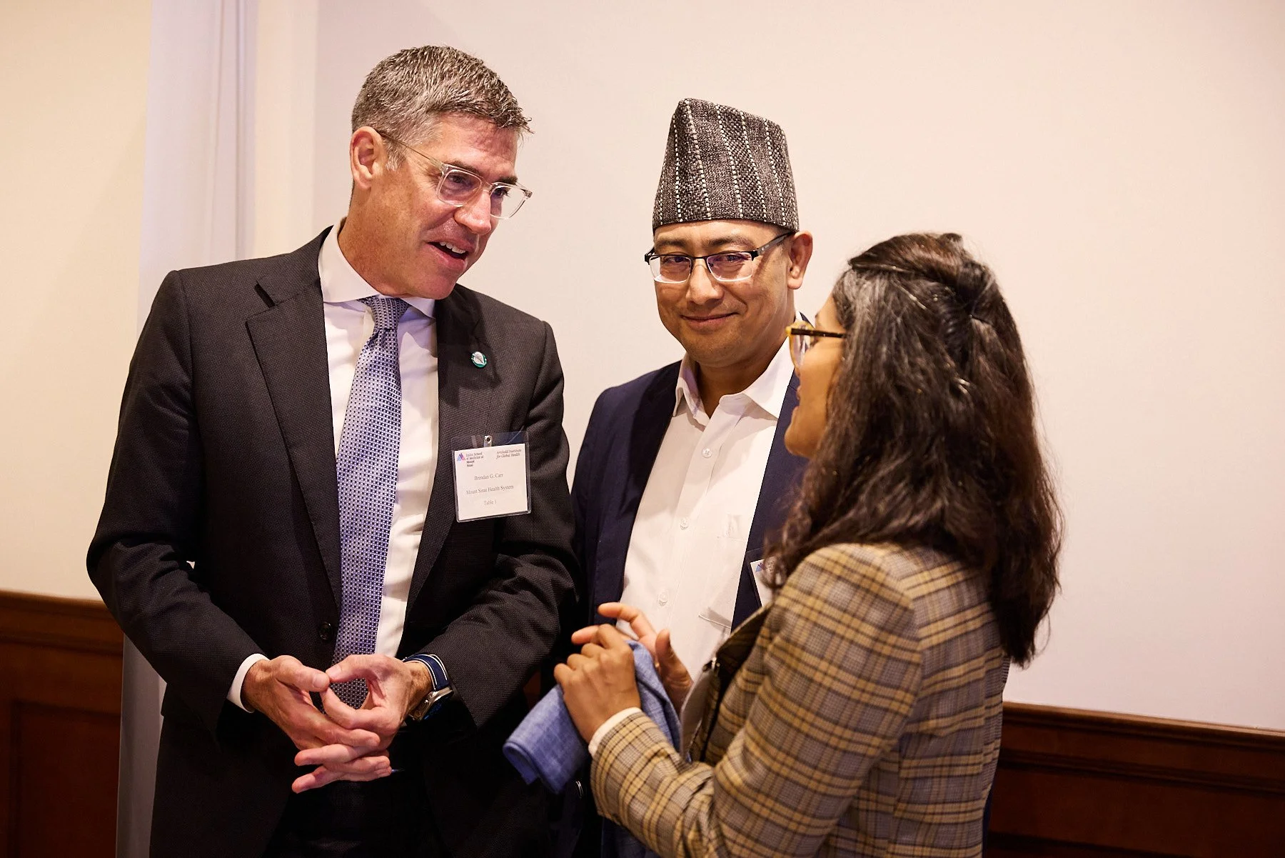 Three people are engaged in conversation at a professional event. The man on the left is wearing glasses, a suit, and a name tag. The man in the middle, also wearing glasses, is dressed in a traditional Nepali hat, suit, and white shirt. The woman on