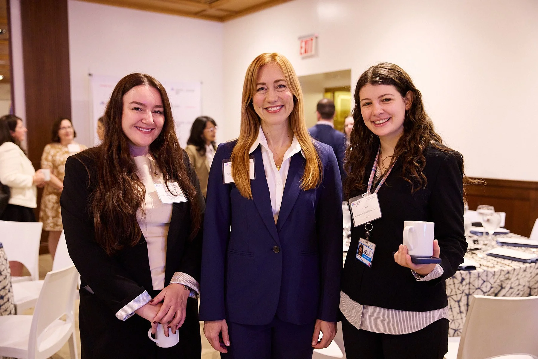 Three women in professional attire smiling at a conference or event, standing in a decorated hall with tables and other attendees in the background.