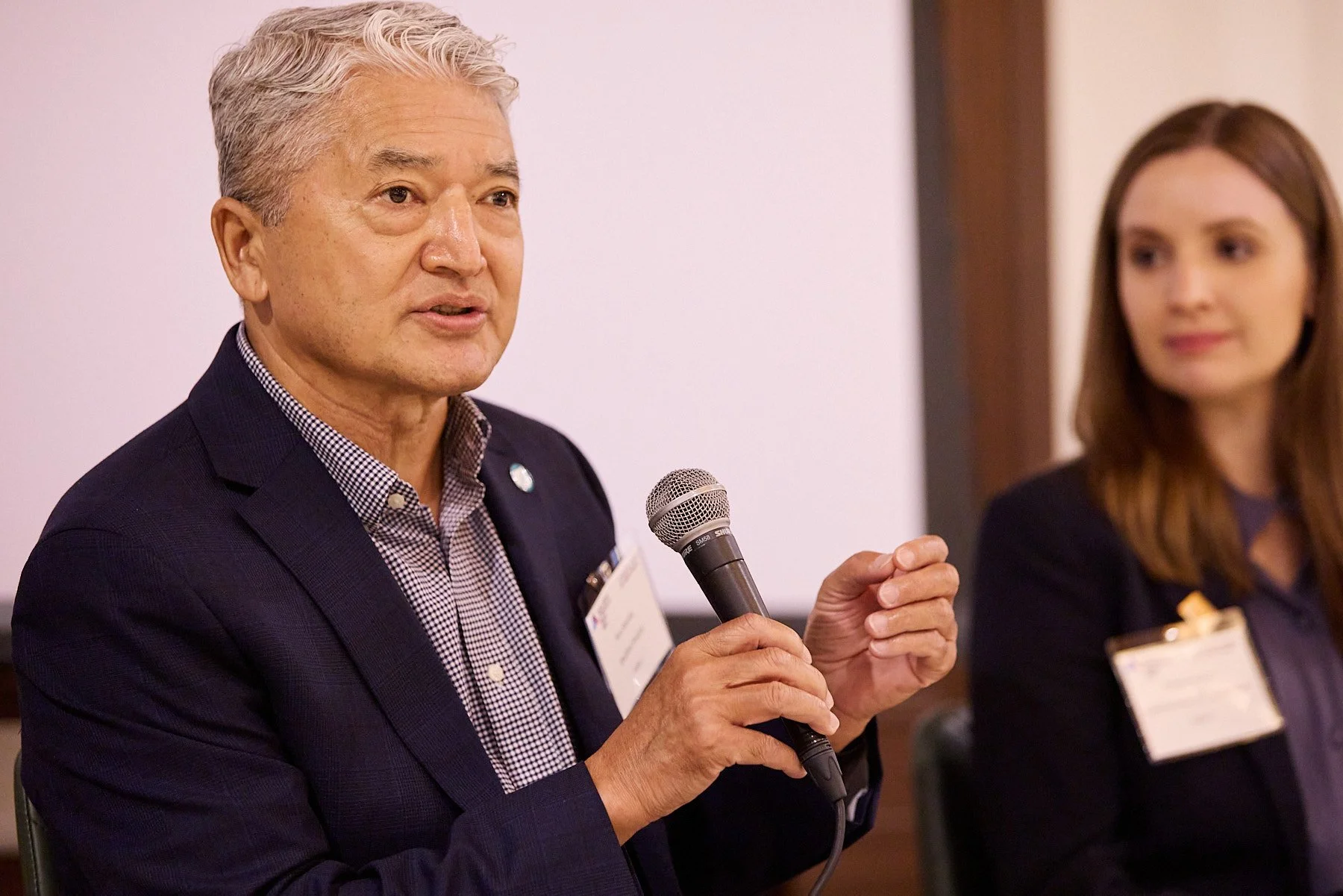 An older man with gray hair speaking into a microphone at a professional event, with a woman with brown hair sitting beside him, both wearing dark suits and name tags.