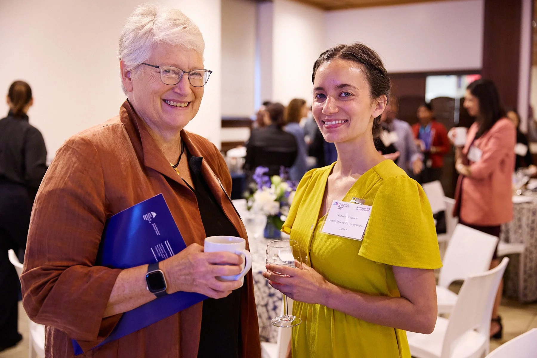 Two women at a professional event or conference, smiling and holding beverages, with other attendees in the background.