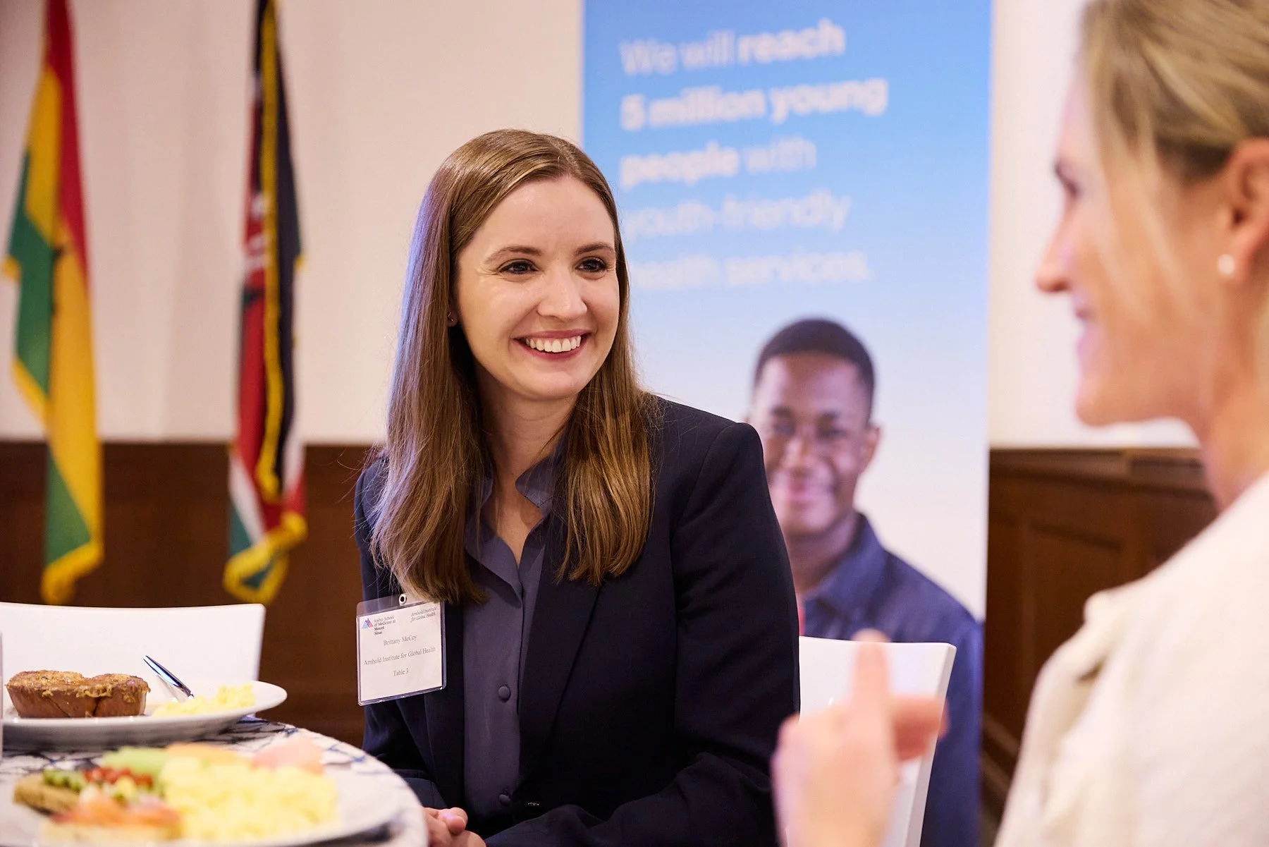 Two women are sitting at a table, smiling and talking to each other during a formal event. The woman on the left has long brown hair, a dark blazer, and a name badge. The woman on the right is blonde, wearing a light-colored outfit, and is gesturing 