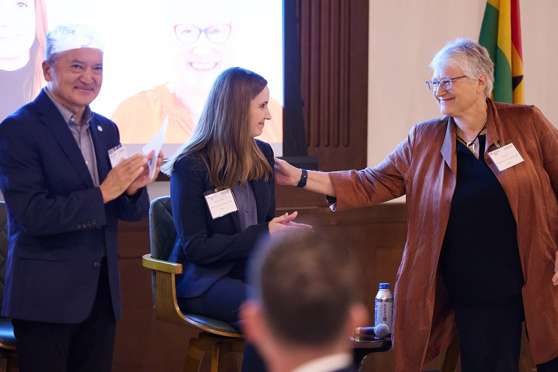 Three people at a professional event; a woman is sitting, receiving a congratulatory gesture from an older woman in a brown jacket, while a man in a dark suit and gray hair stands nearby smiling.