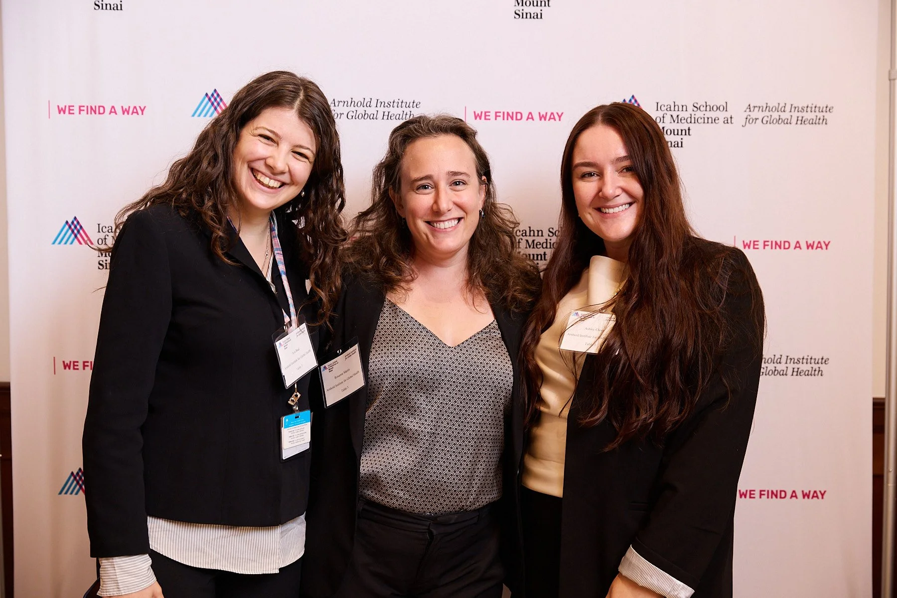 Three women smiling and posing together at an event, standing in front of a white backdrop with logos and text related to the Icahn School of Medicine at Mount Sinai and the Arnold Institute for Global Health, wearing conference badges.