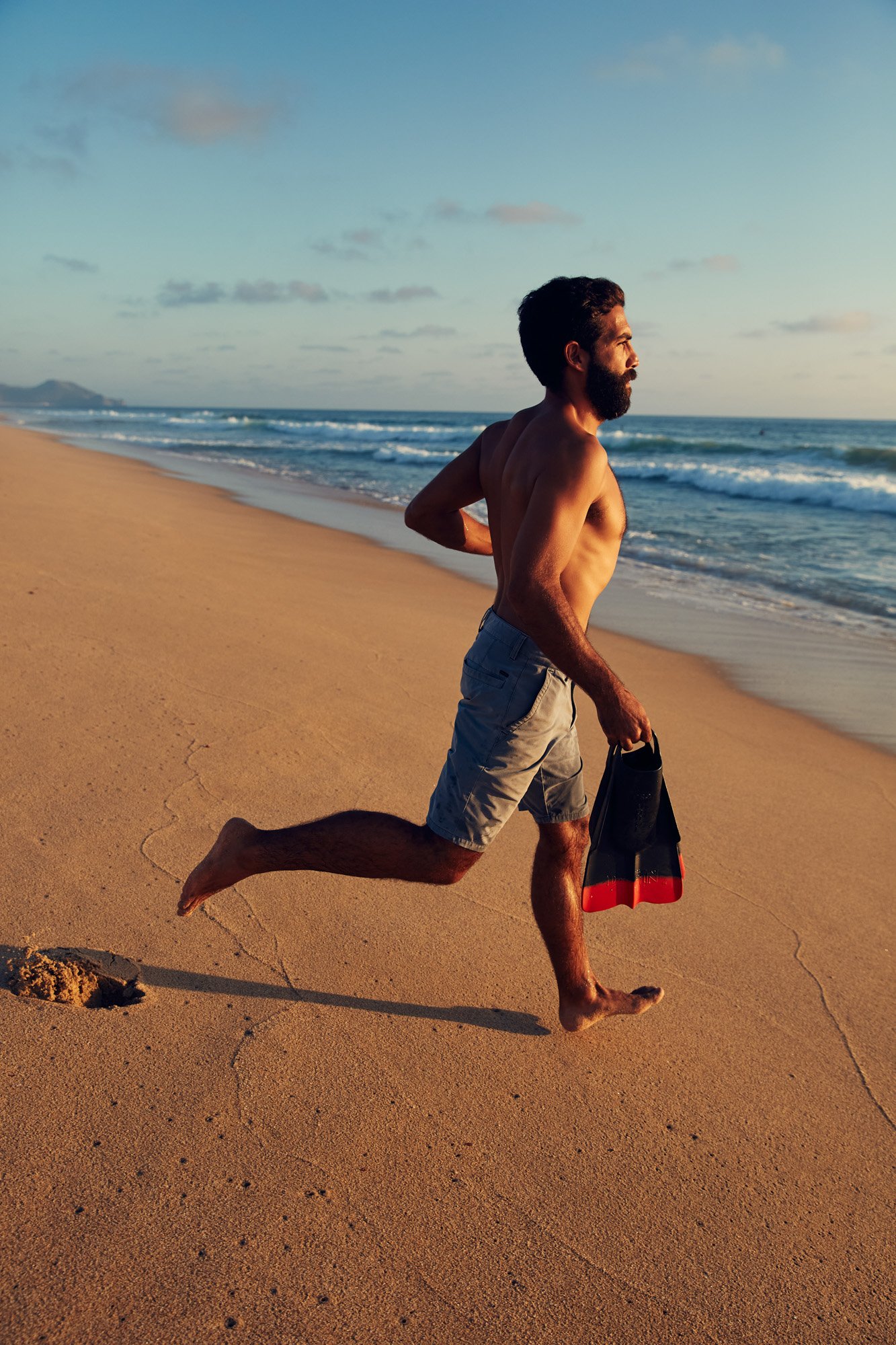 A bearded man running on the beach with snorkeling fins in his hand during sunset near Todos Santos in Baja Mexico.