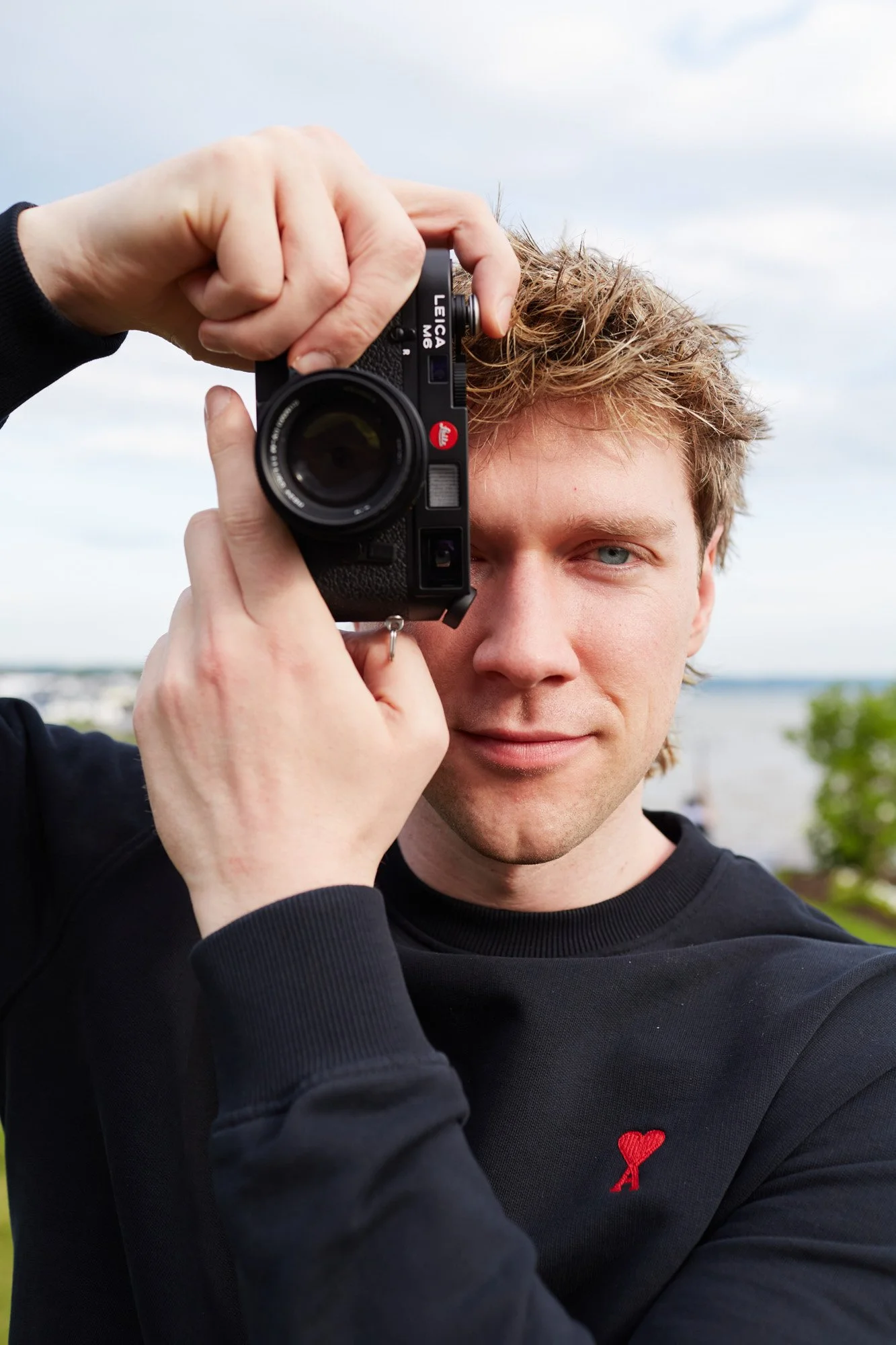 Collins Key, holding a black Leica camera up to his face, with a slight smile, wearing a black sweatshirt with a small red heart pin on the chest, background shows a cloudy sky and some greenery.