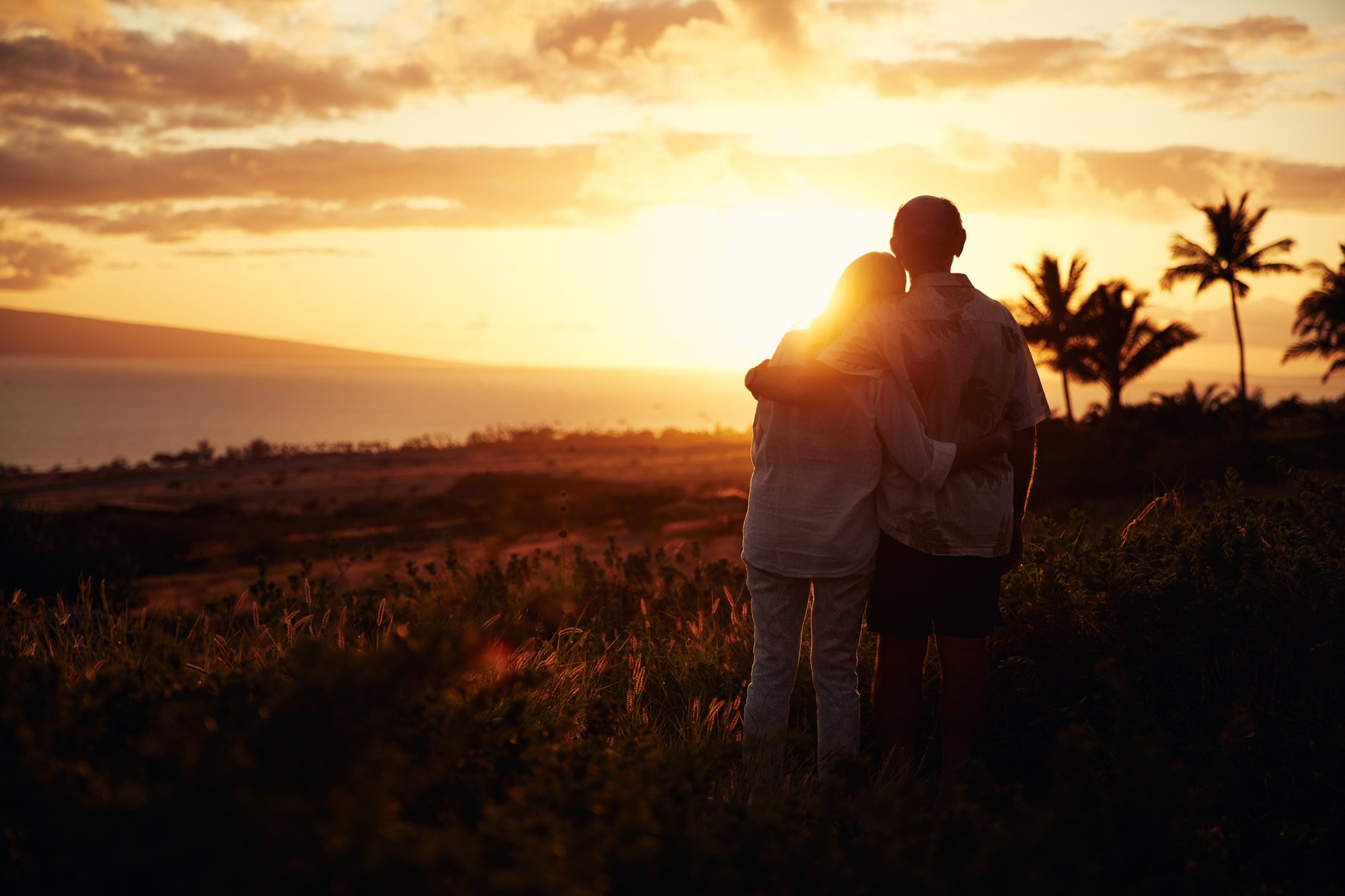 A retired couple standing together in Lahaina Maui, watching a sunset with palm trees in the background.