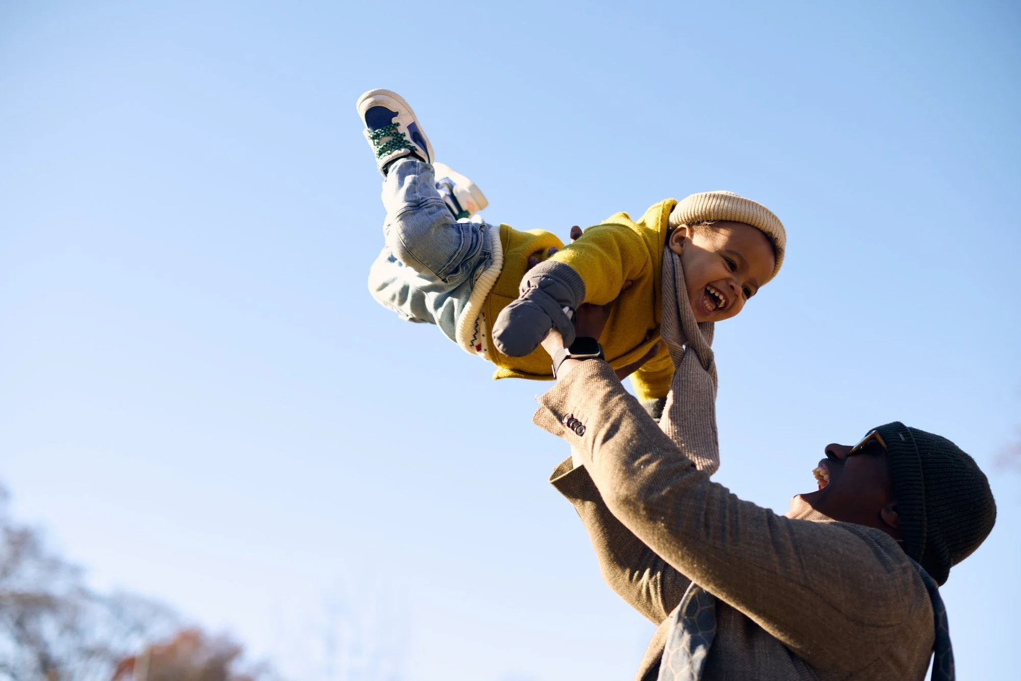 Father raising his child in the air outdoors during winter, both smiling and enjoying the moment with a clear blue sky in the background.