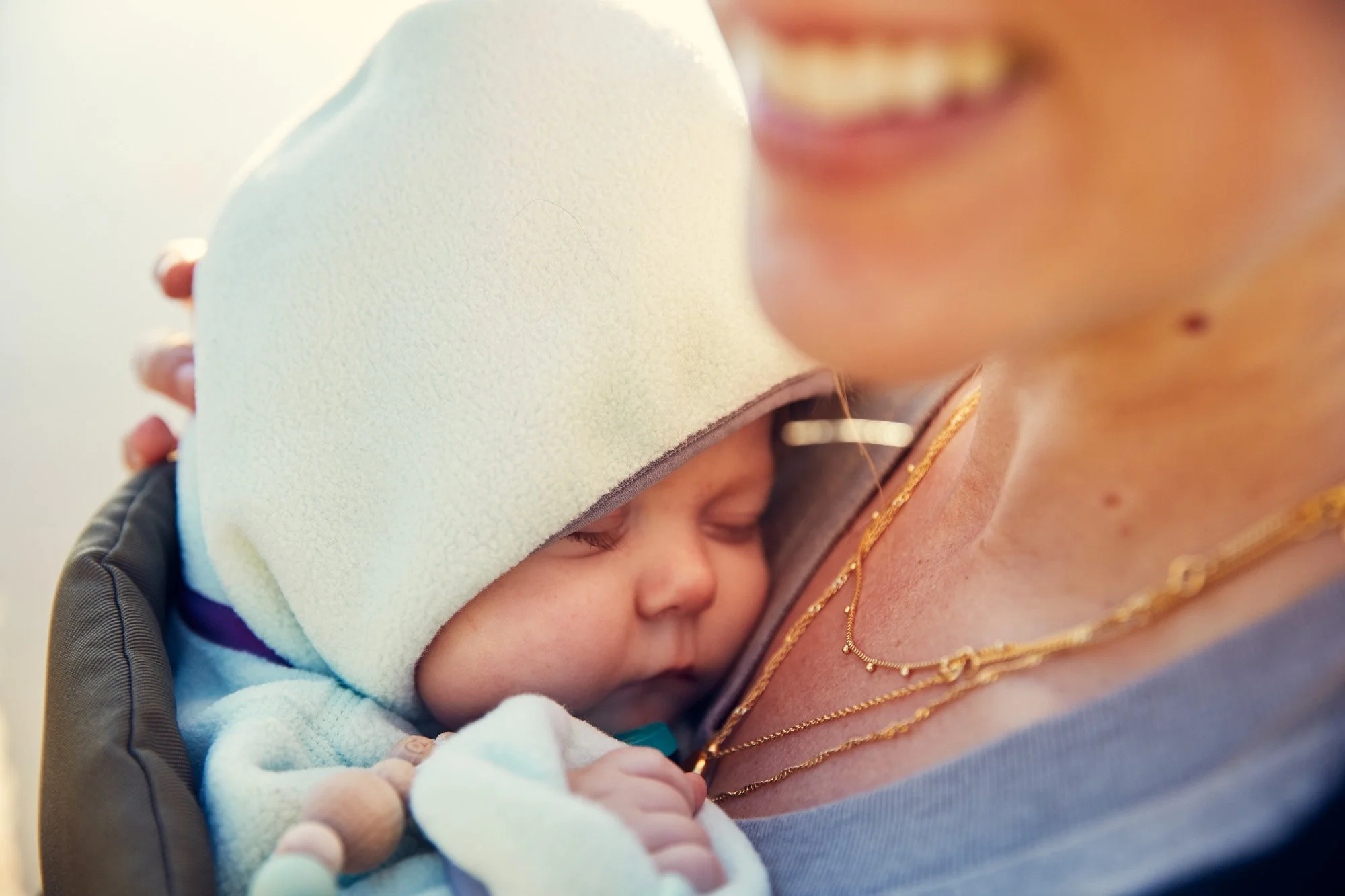 Close-up of a baby sleeping on a woman's shoulder, with only part of her face visible, smiling, and wearing gold jewelry.