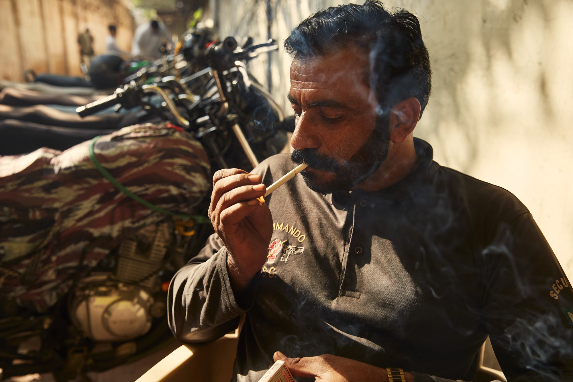 A man with black hair and a beard smokes a cigarette indoors, with motorcycles visible in the background. Karachi Pakistan