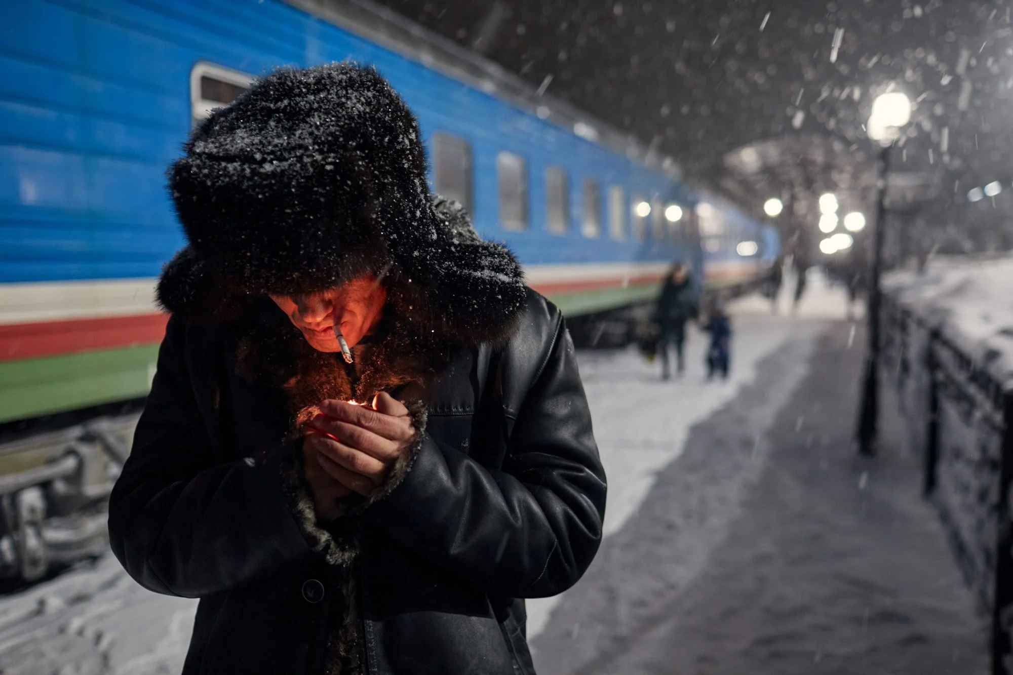 A man smoking a cigarette at a train station during snowy night, wearing a black fur hat and leather jacket, with a blue train in the background. From the Amur-Yakutsk Mainline (Permafrost Express), in Sakha Republic Siberia Russia