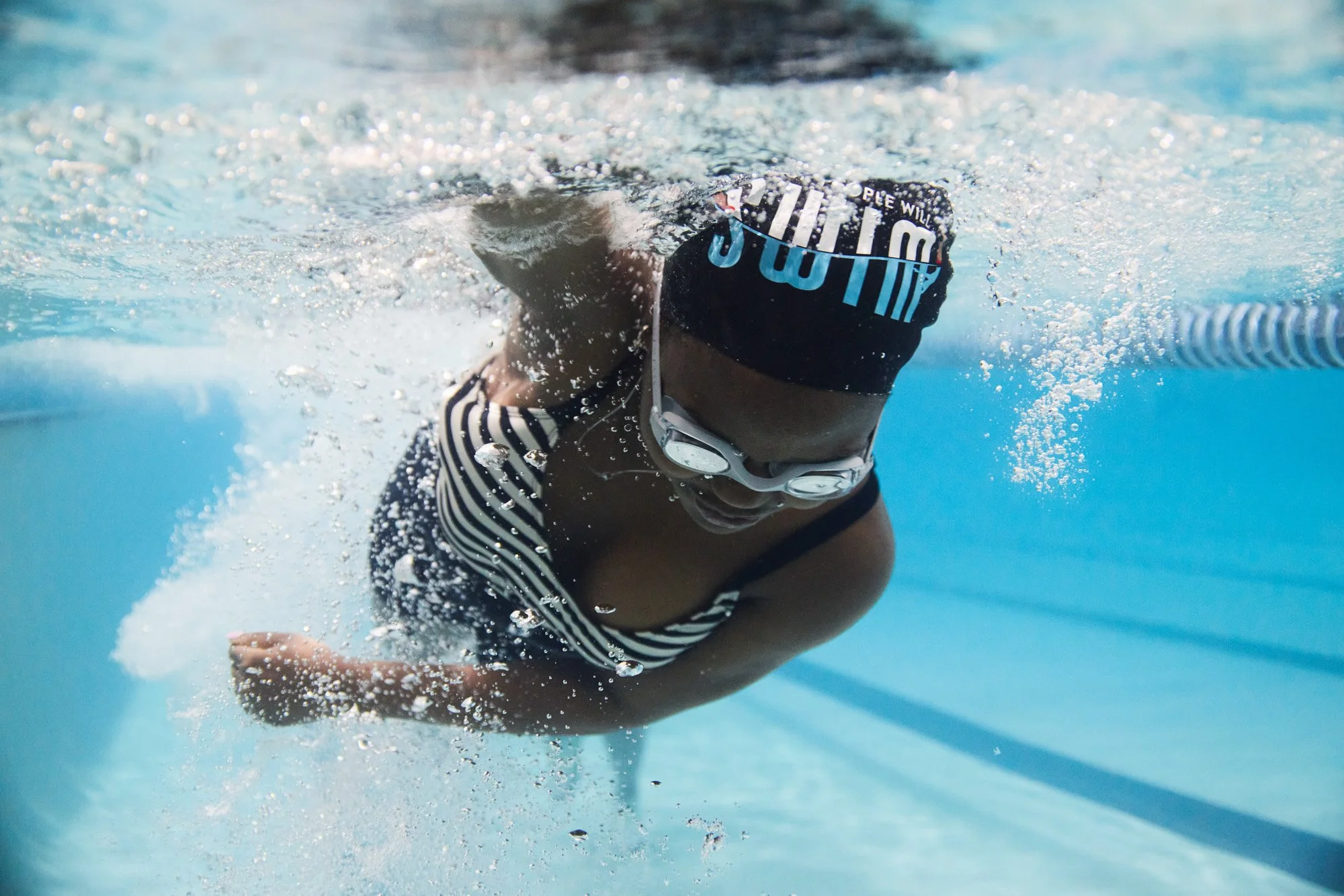 Black female swimmer in a pool, wearing goggles and a black swim cap, swimming freestyle stroke.