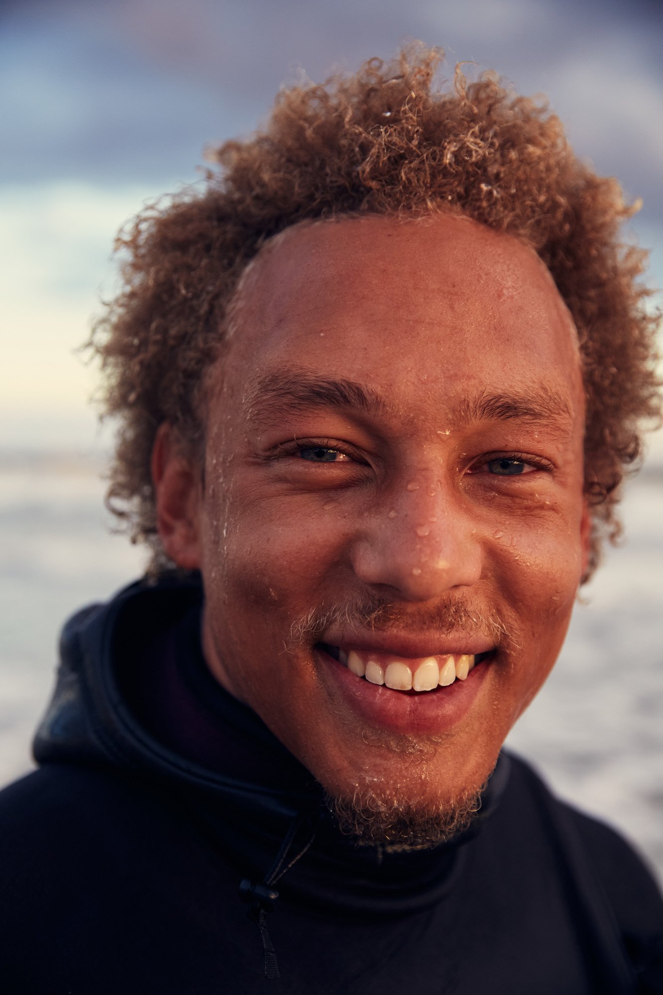 Close-up of smiling young man with curly blond hair and mixed skin tone, water droplets on face, and ocean in the background.