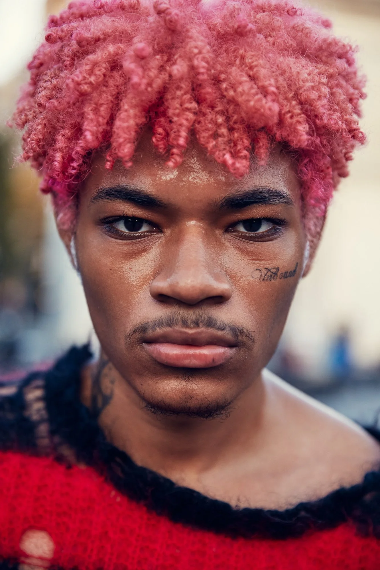 A young man with pink curly hair, facial tattoos reading "Vii and" on his right cheek, and a serious expression wearing a distressed red sweater, in Washington Square Park, Manhattan New York City