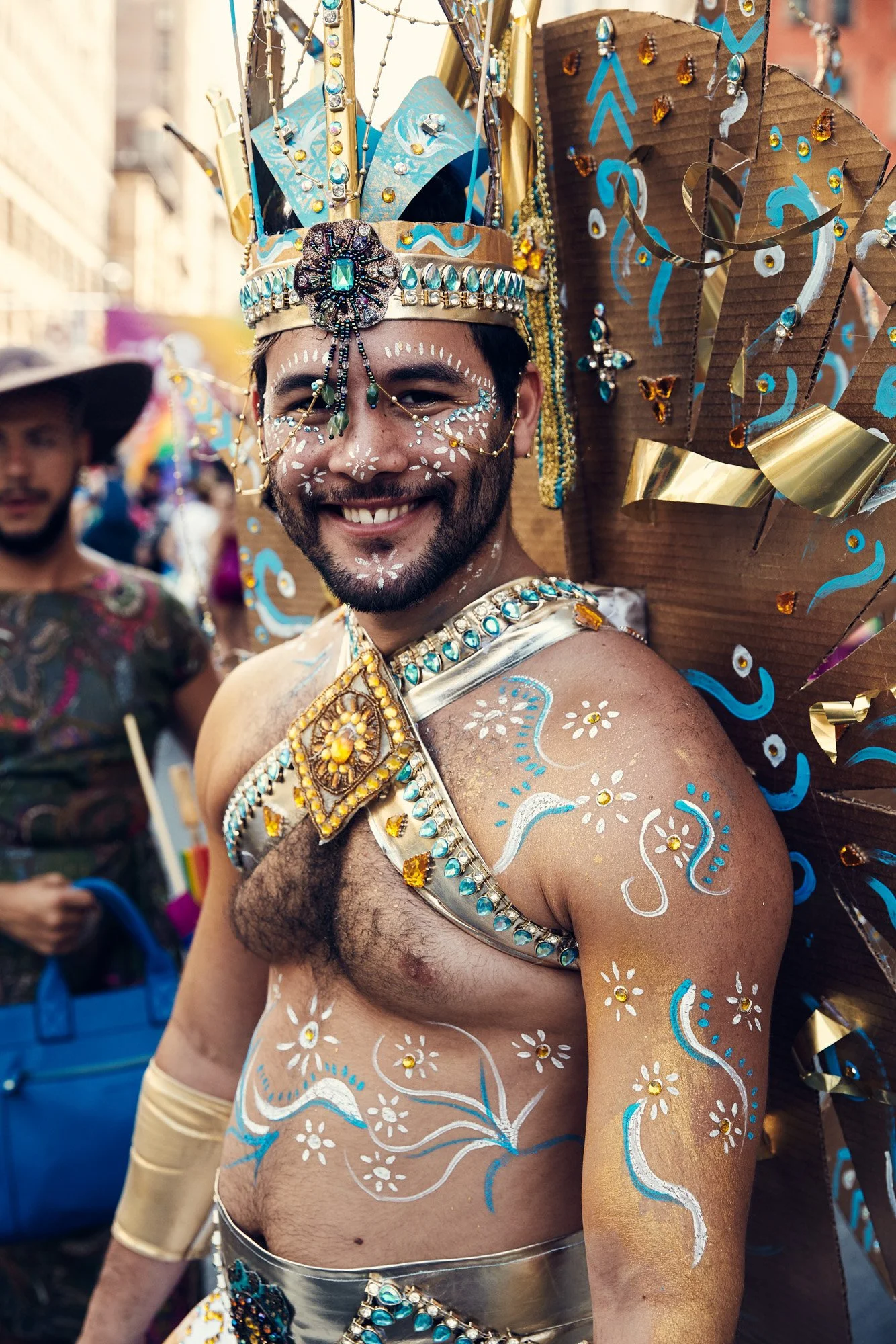 A person smiling at NYC pride parade, dressed in a colorful costume with a large, decorated headpiece, body paint, and jewelry, surrounded by other festival-goers.