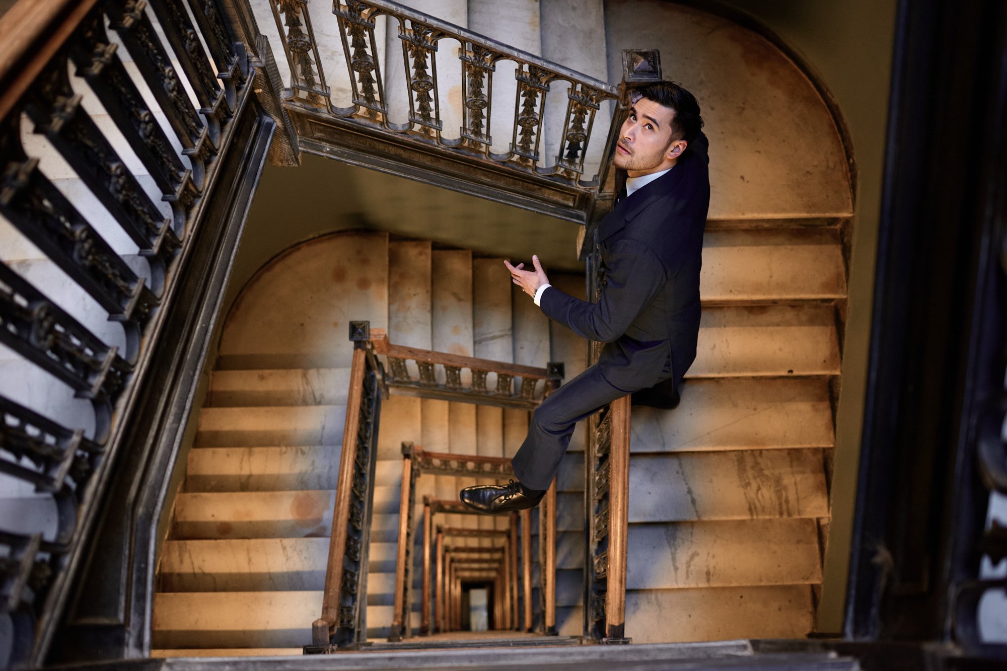 Magician Dan White in a black suit sitting on a spiral staircase, looking up, taken from above.