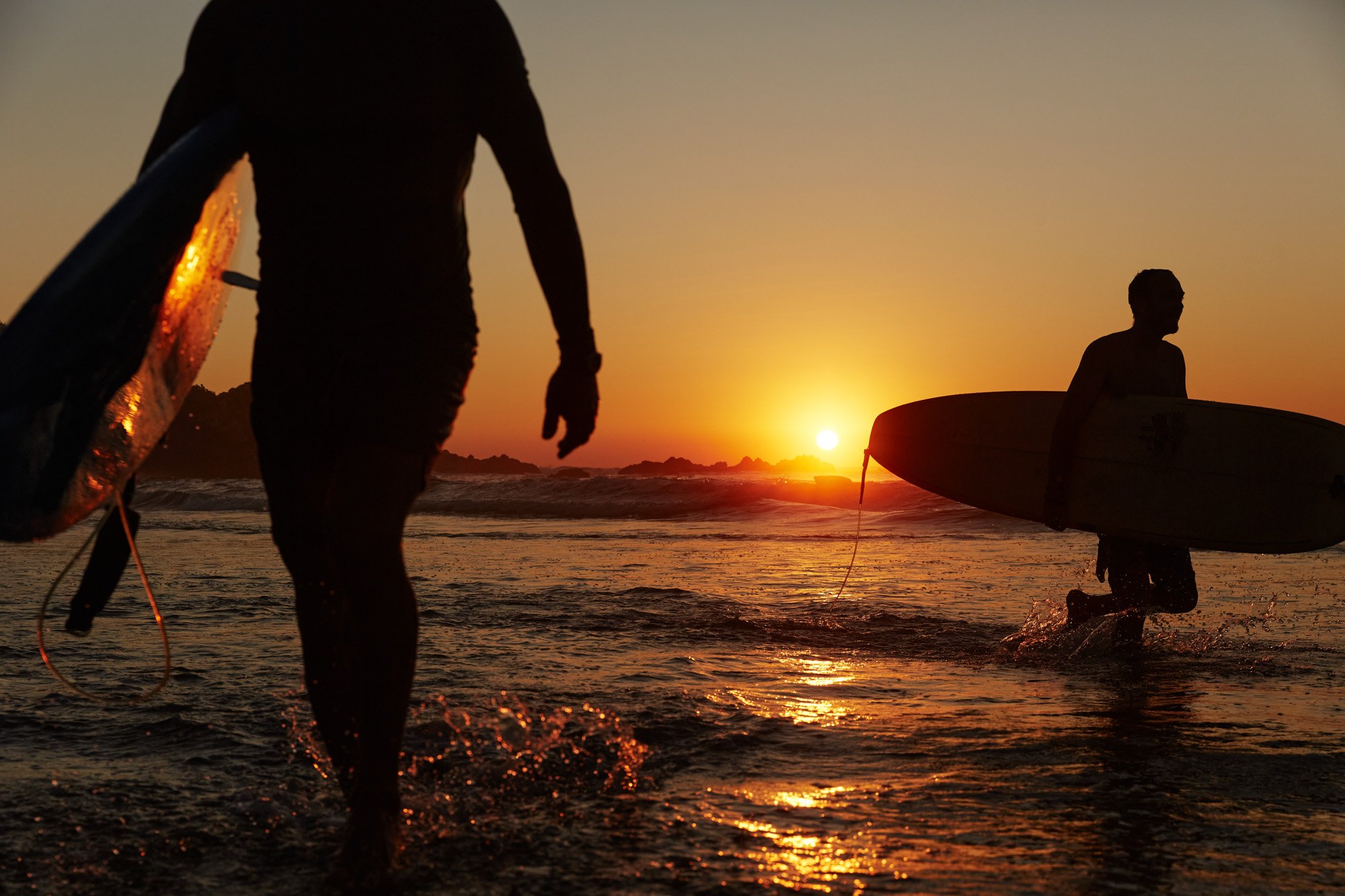 Two surfers with surfboards at La Punta Puerto Escondido Oaxaca Mexico, entering the ocean during sunset, with the sun low on the horizon and creating a silhouette effect.