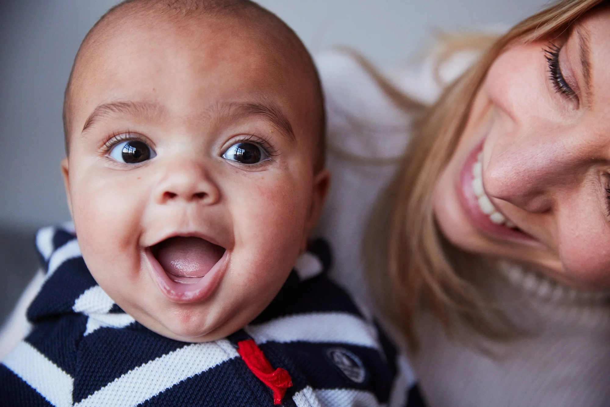 A smiling baby with big eyes lying next to a woman with long hair and a big smile.