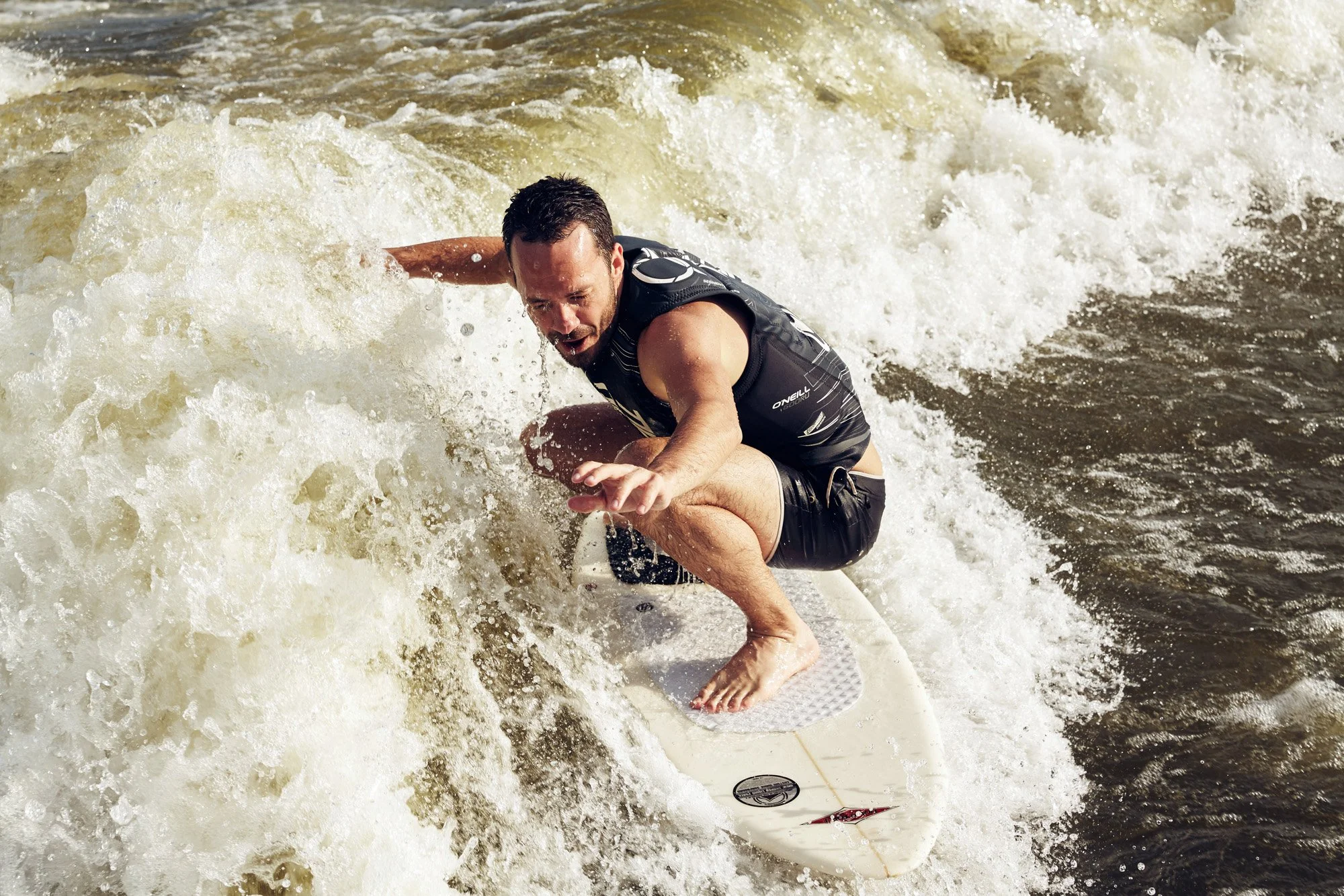 A man wake surfing on a wave, crouching on a white surfboard with a black traction pad, wearing a black sleeveless wetsuit, with a focused expression.