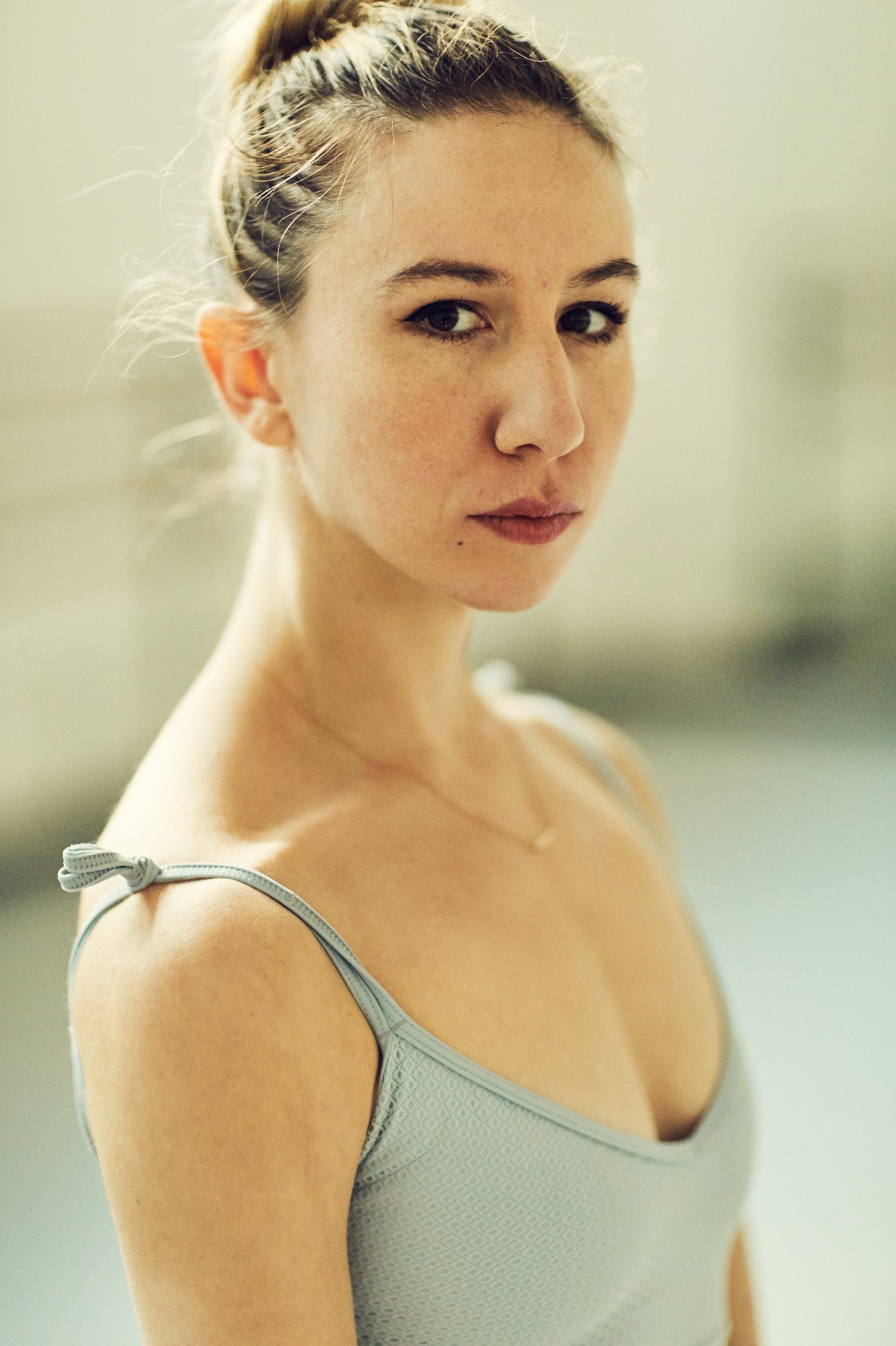 A ballerina Isabella Boyleston with light skin, wearing a light gray tank top with tied straps, standing indoors with a soft background, looking at the camera.