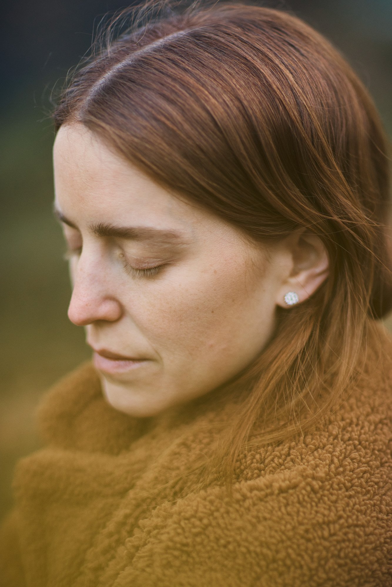 Close-up of Ana Corina Sosa, with reddish-brown hair and fair skin, wearing a brown textured coat and pearl stud earrings, with her eyes closed, outdoors. Central Park Manhattan New York City