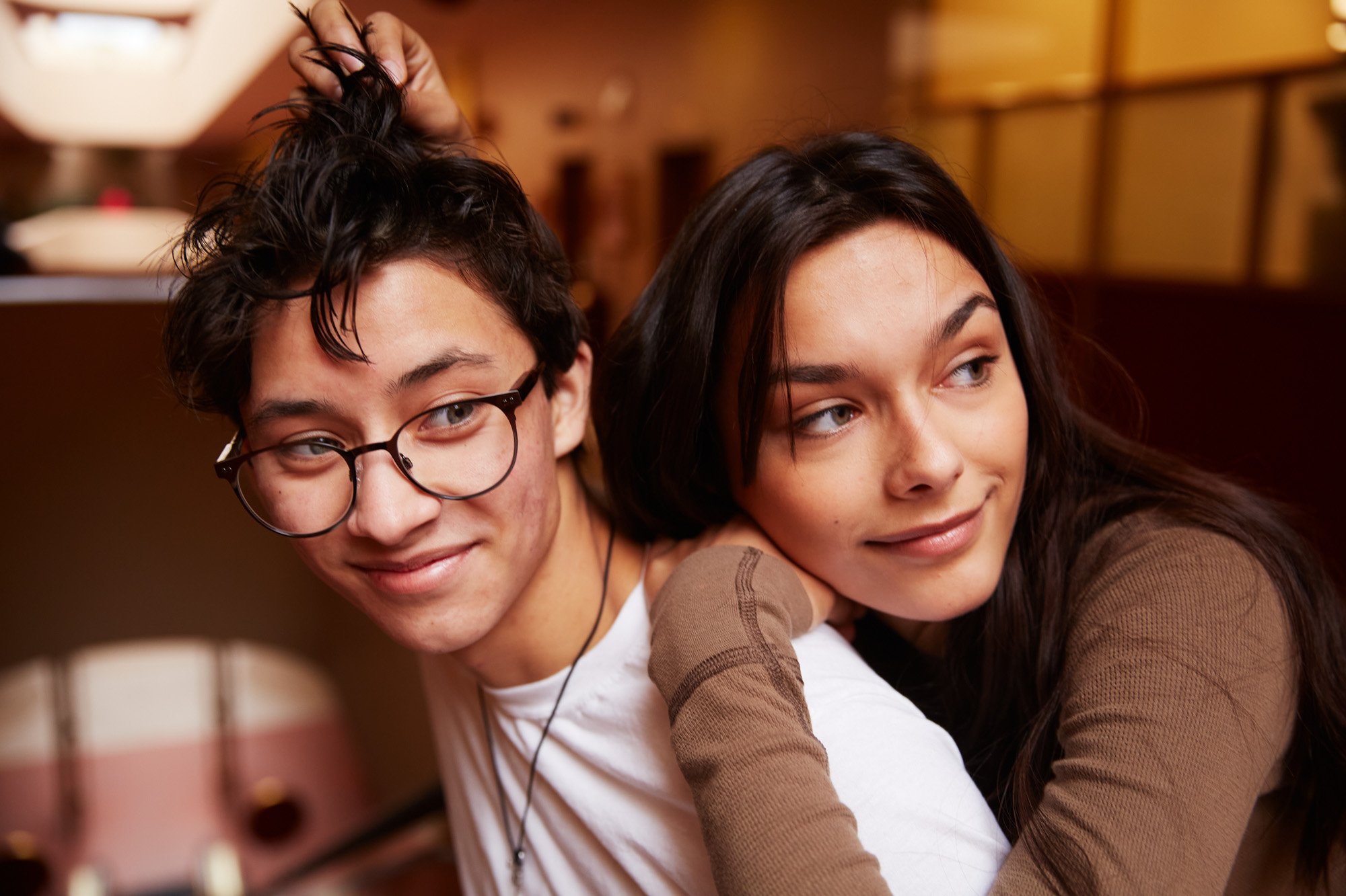 A young man with glasses and a young woman Violet Lux with long dark hair pose closely together, smiling softly in a cozy indoor setting.