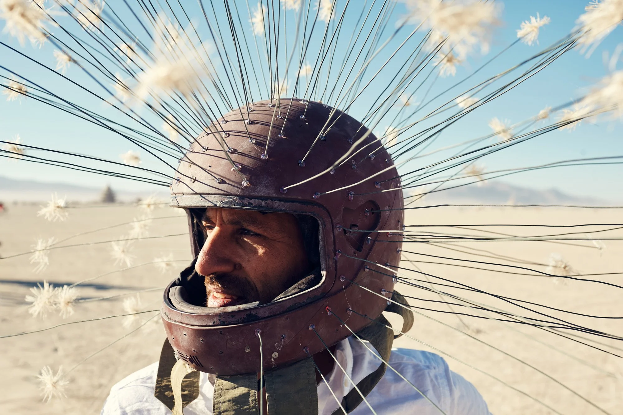 A man wearing an open motorcycle helmet with numerous wires and white fluffy pom-poms attached all over, set in a desert landscape with mountains in the background. Burning Man