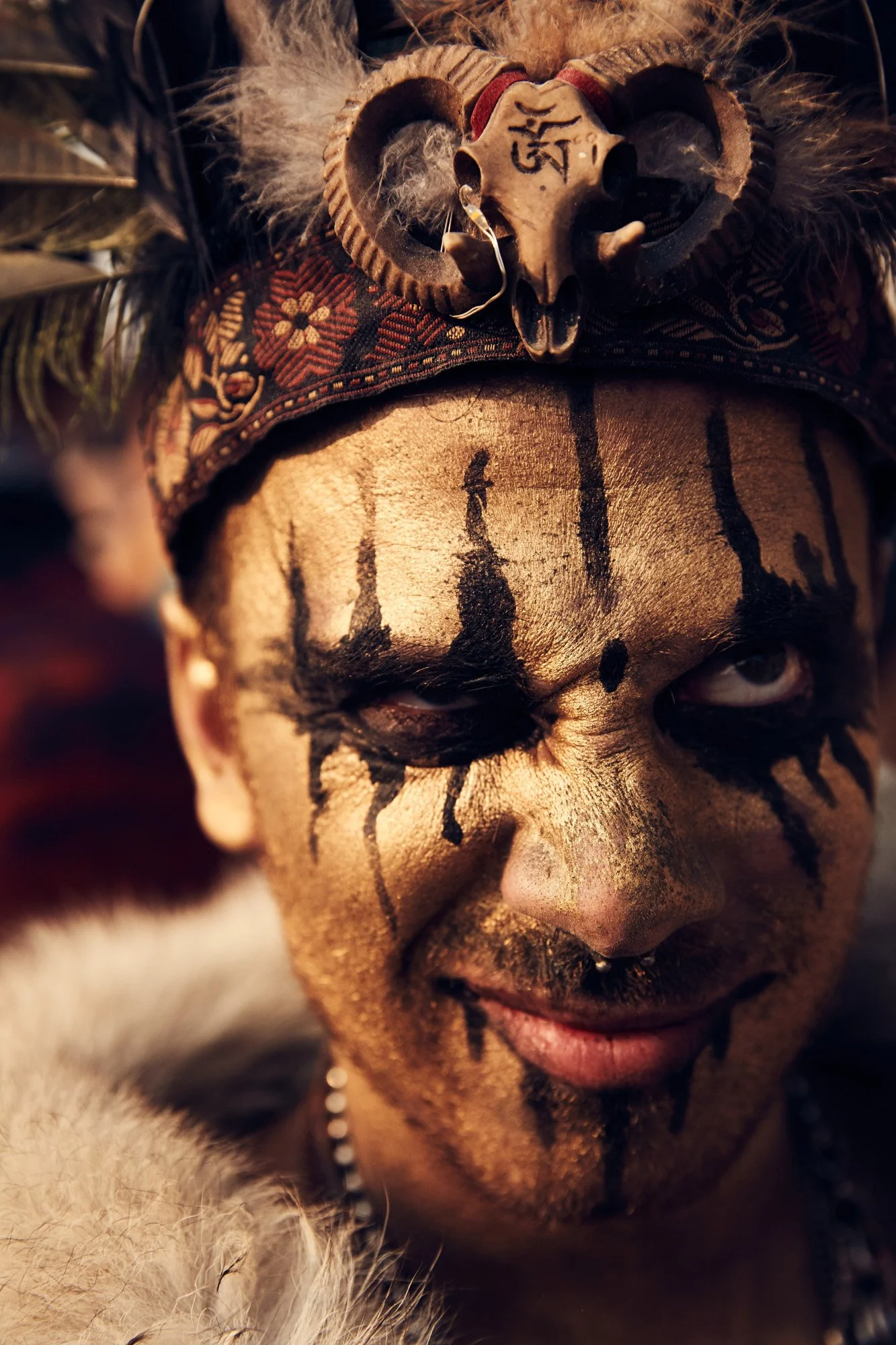Person with gold face paint and black tribal designs, wearing a headdress with a carved animal skull and feathers. Burning Man