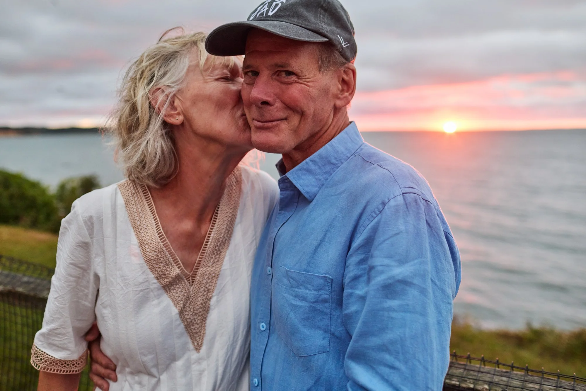 An older woman kisses an older man on the cheek as they stand outdoors near the water at sunset in Montauk, with the sun setting over the horizon.
