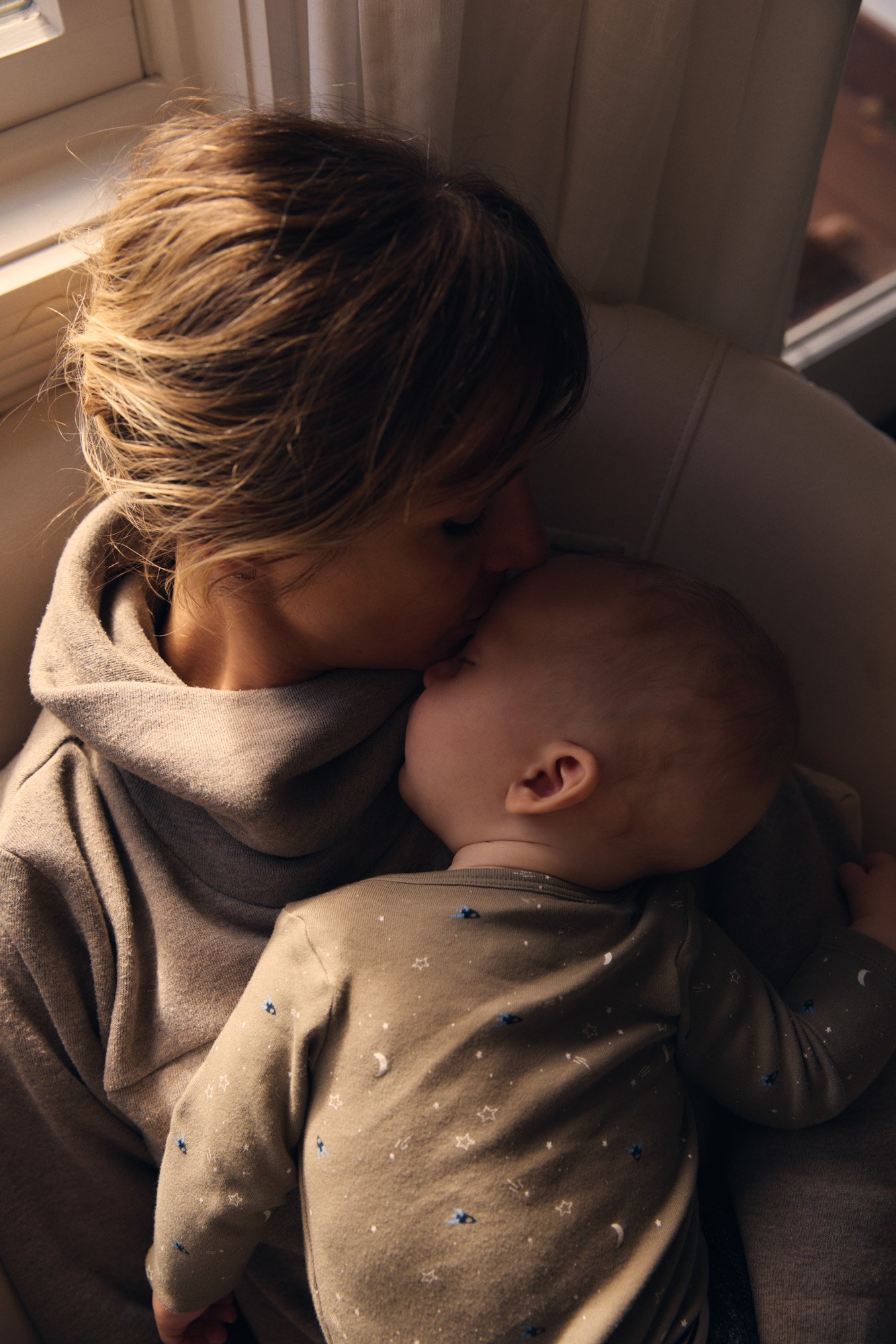 A woman in a gray hoodie gently kisses a sleeping child on the forehead in a cozy indoor setting.