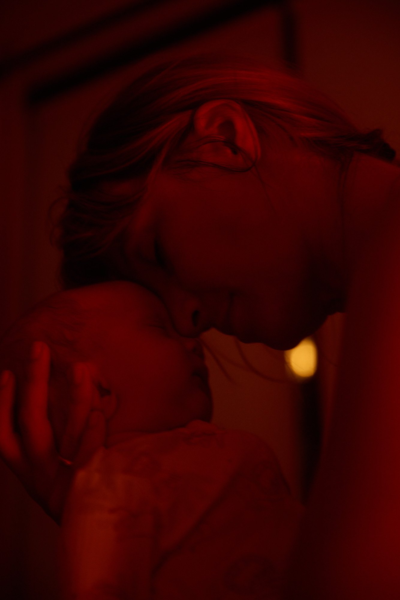 A woman with blonde hair holding a baby in a dimly lit, red-toned environment, close-up shot