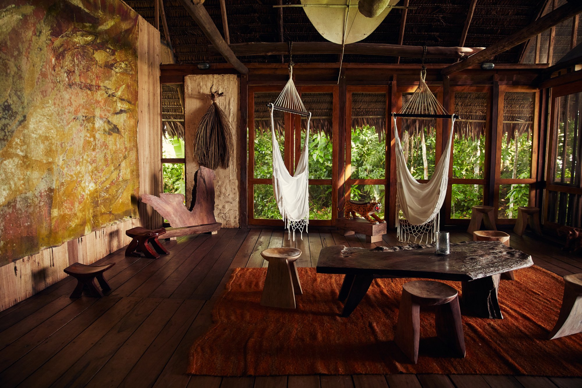 Interior of a rustic wooden hut with large windows showing lush greenery outside, with a carved wooden machine, hanging hammocks, a large irregular wooden table, and wooden stools and benches on a reddish-brown rug. Calanoa Resort, Amazonas, Colombia