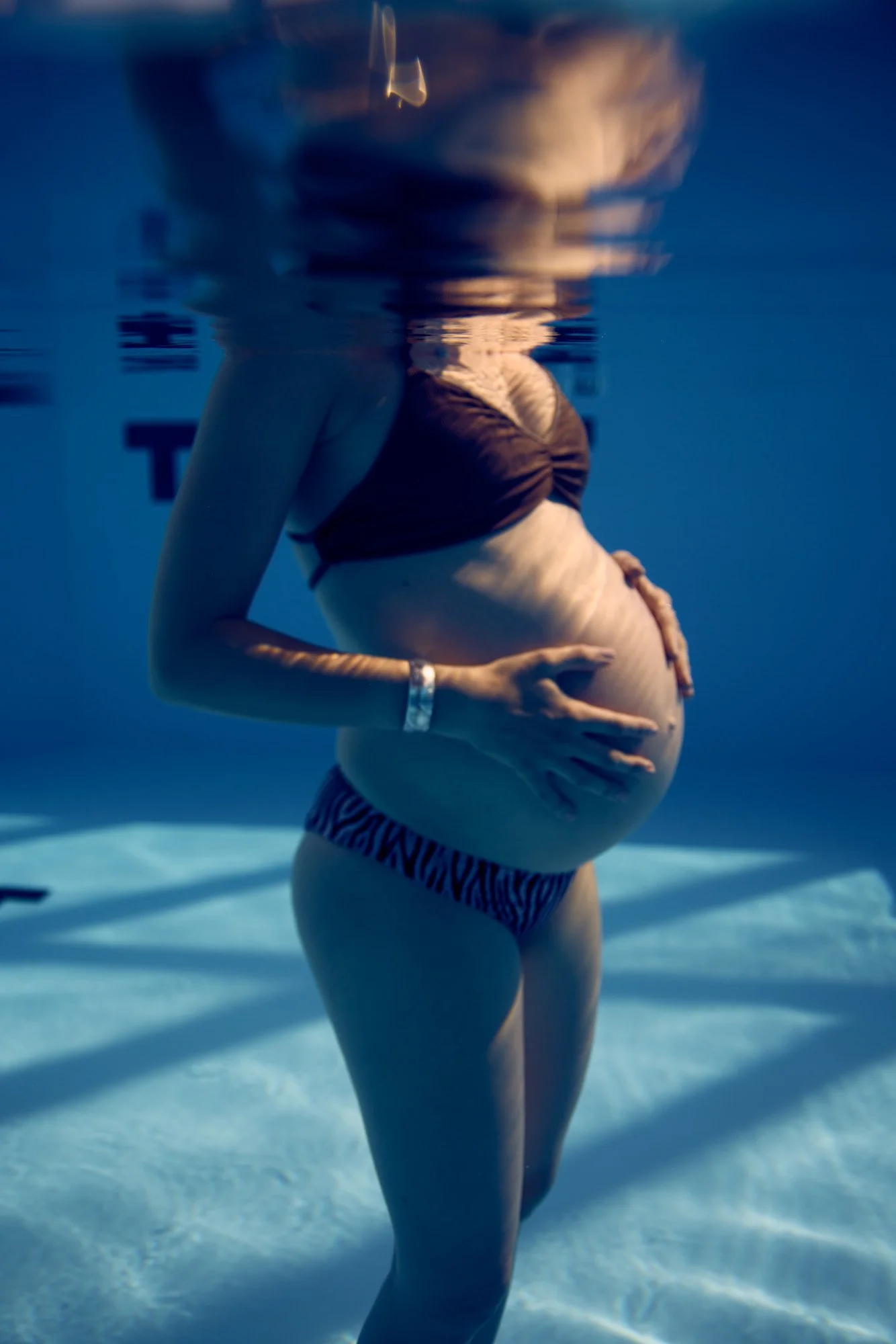 A pregnant woman is underwater in a swimming pool, holding her belly with one hand and the other hand resting on her abdomen, wearing a bikini.