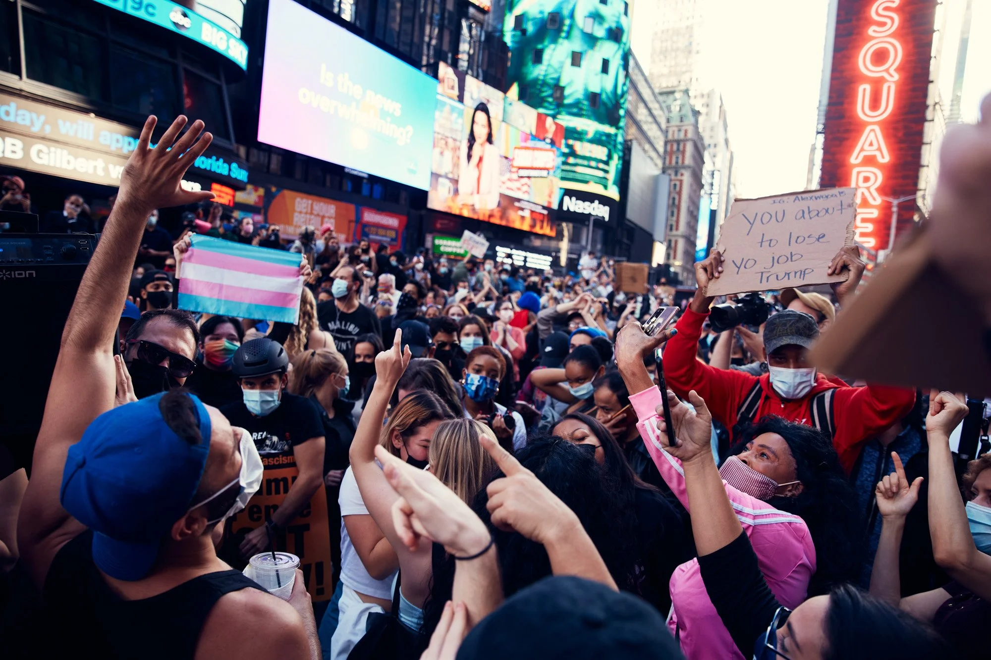 Crowd of protesters in Times Square, New York City, with people holding signs including one that says 'you about to lose your job Trump' and a transgender pride flag, many wearing masks amidst digital billboards and bright city lights.