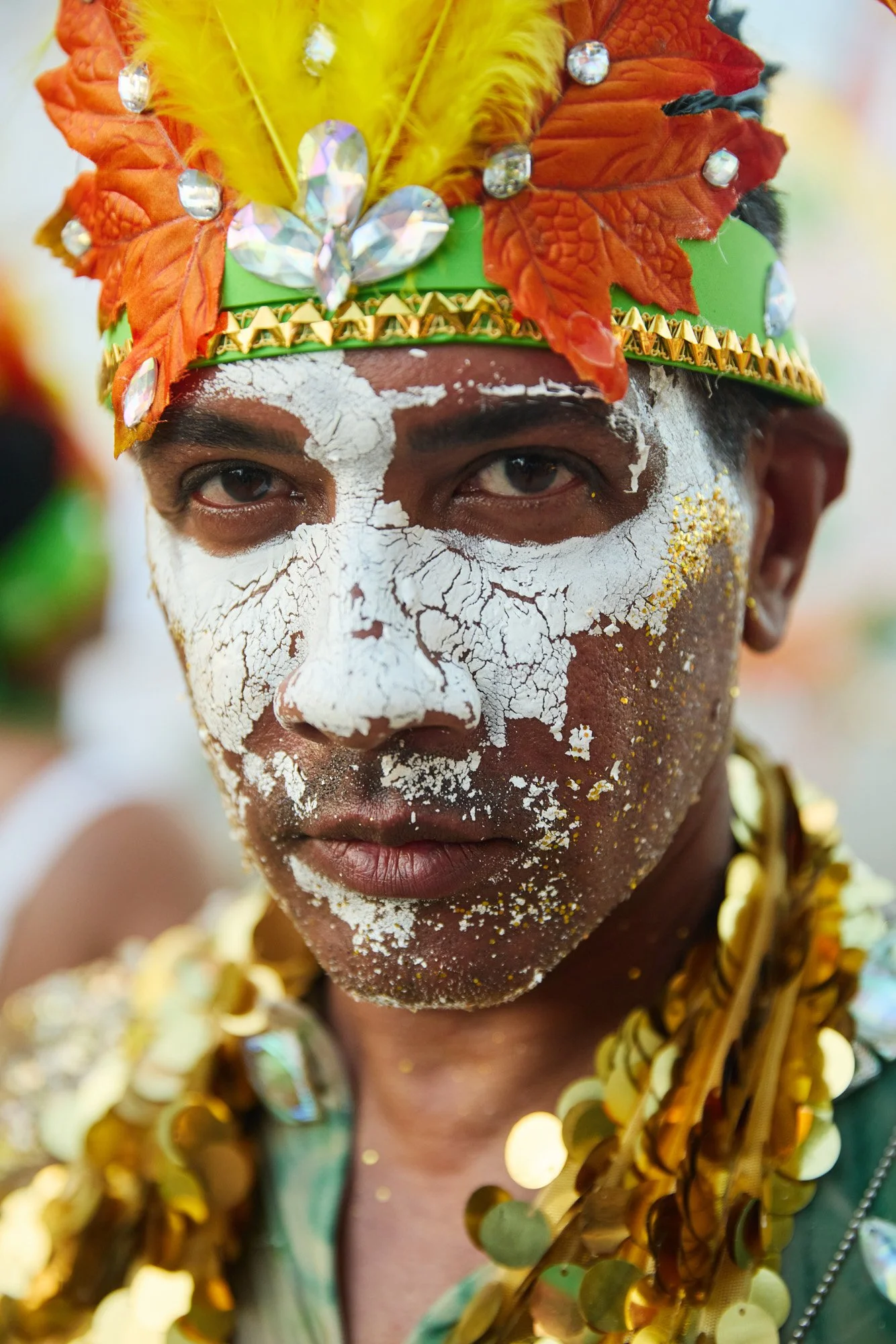 A person with a painted white face, wearing a colorful headdress decorated with feathers, beads, and large rhinestones. The individual is surrounded by other people in similar attire, as part of Trinidad's Mardi Gras event