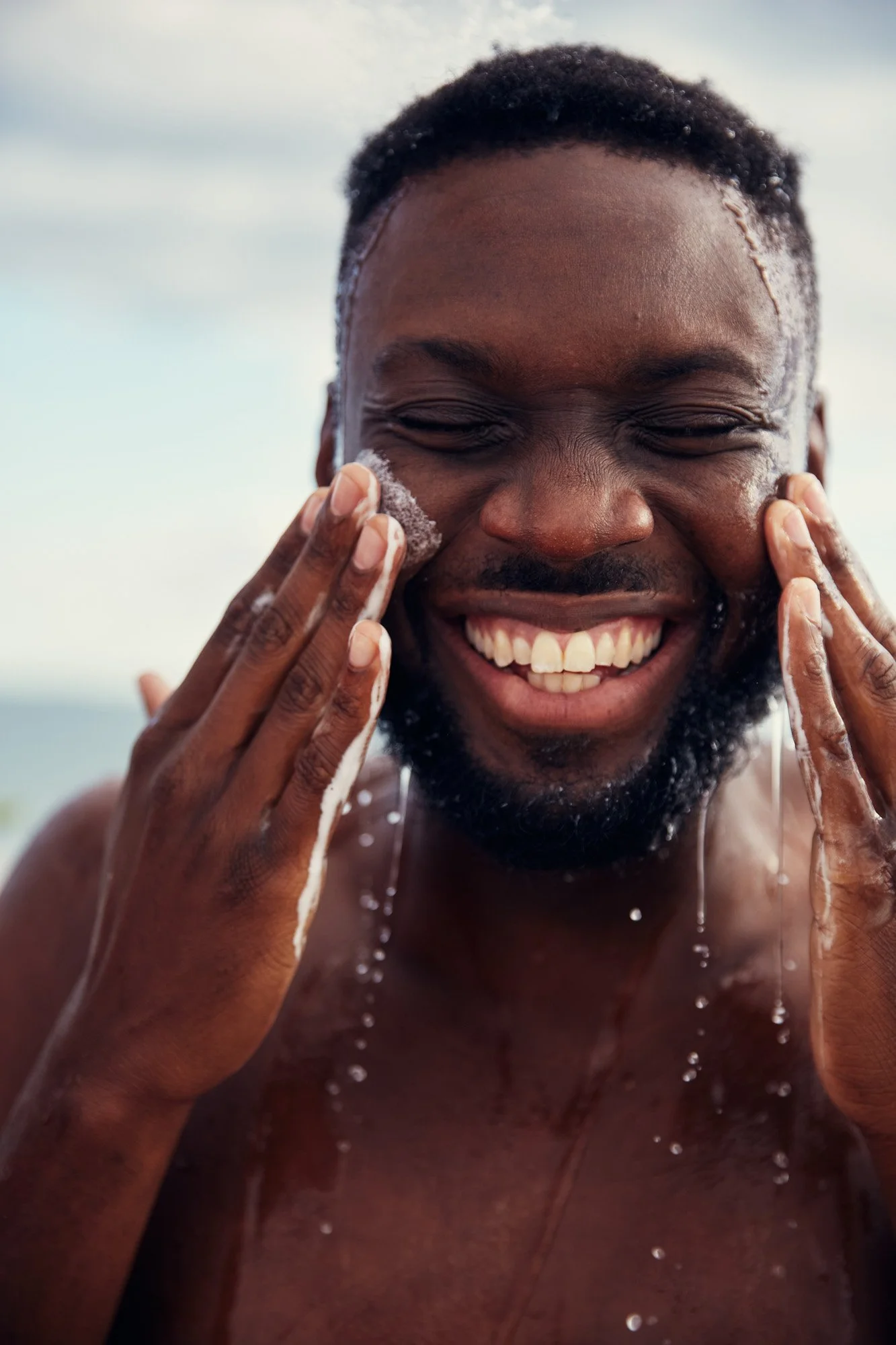 A young Black man with dark skin washing his face with soap and water on a beach, smiling with his eyes closed. Rockaway Beach New York City