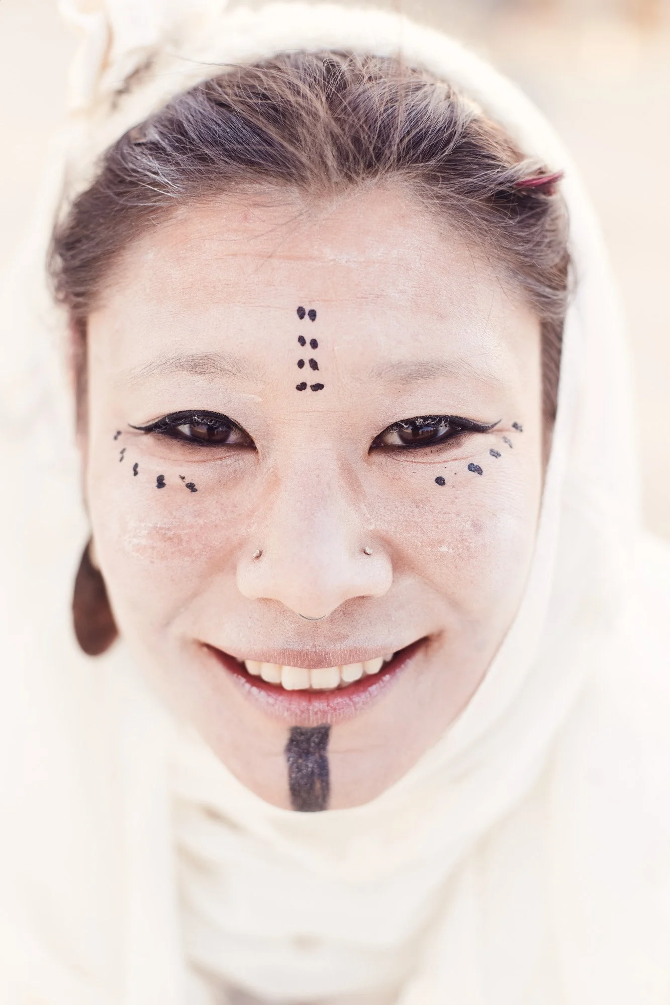A woman with artistic black face paint, including dots on her forehead and cheeks, a vertical line on her chin, and a nose piercing, smiling at the camera at Burning Man