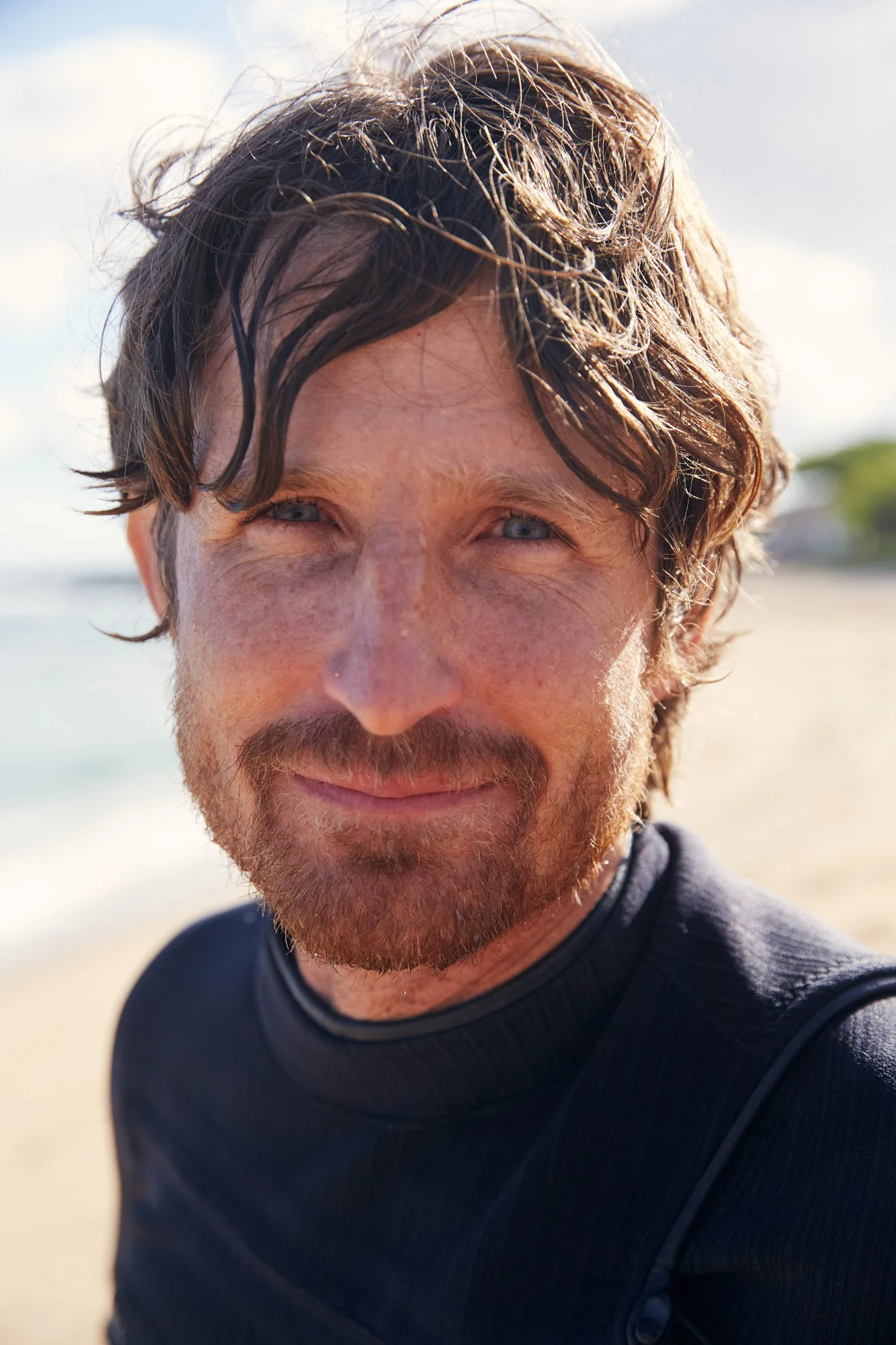 Close-up of a smiling man with light skin, wavy brown hair, and blue eyes at the beach, wearing a black wetsuit top on the north shore of Oahu Hawaii