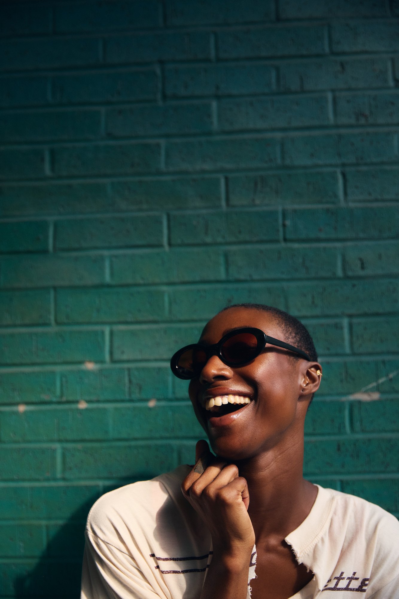 A woman wearing sunglasses smiling and laughing against a green brick wall.