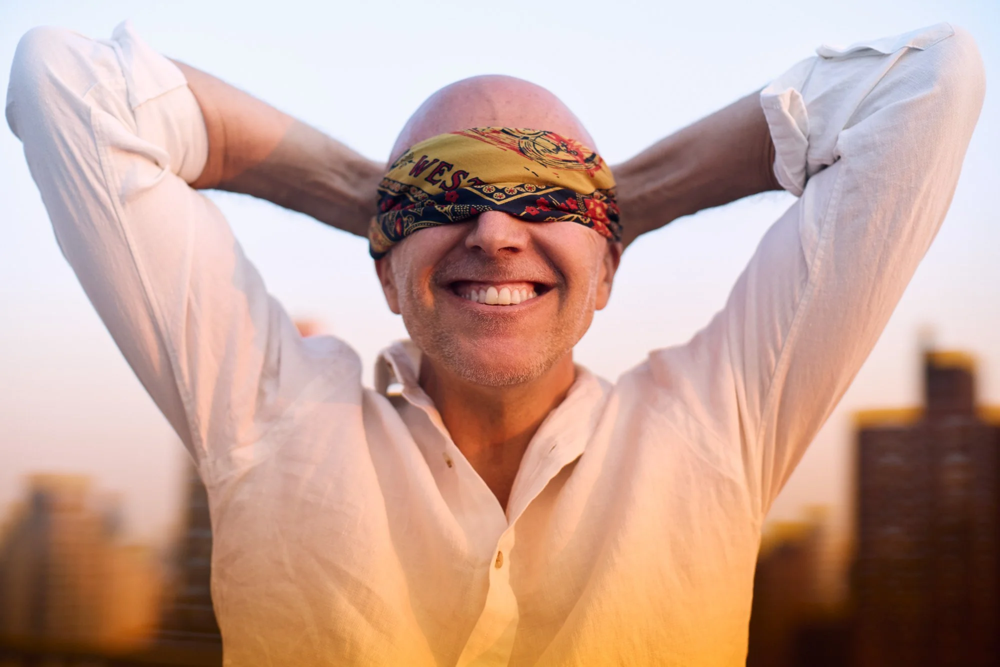 Lee Burridge smiling with a blindfold on, standing outdoors with New York City in the background.