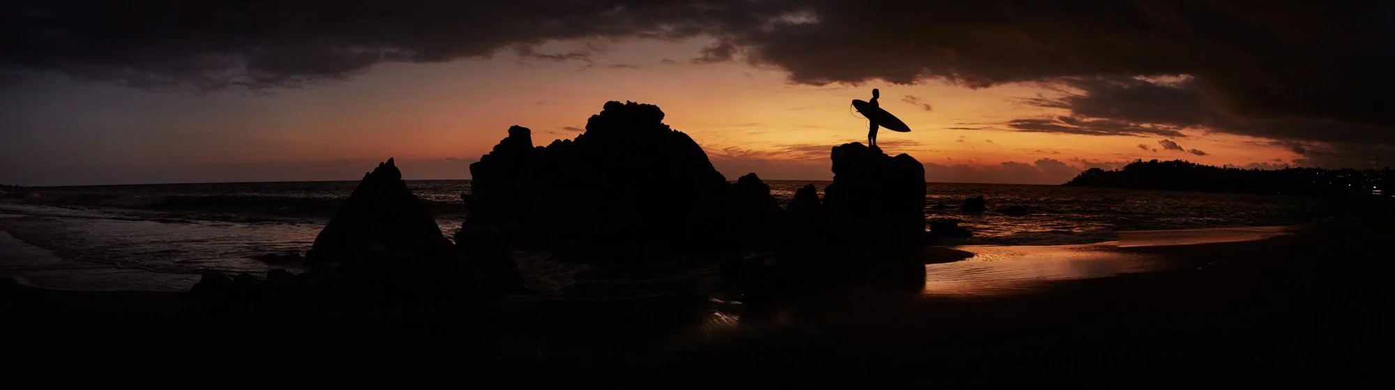Silhouette of a person holding a surfboard while standing on a large rock at the beach during sunset on Zicatela Beach, Puerto Escondido, Oaxaca, Mexico.