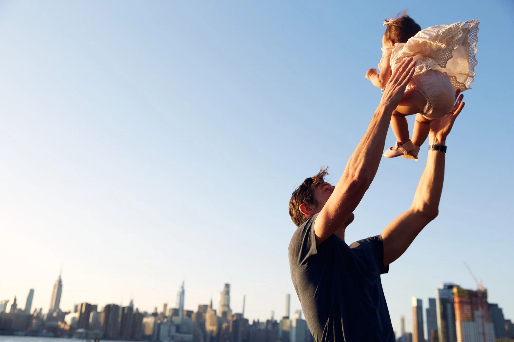 A man lifting a baby girl into the air with the New York City skyline in the background during sunset